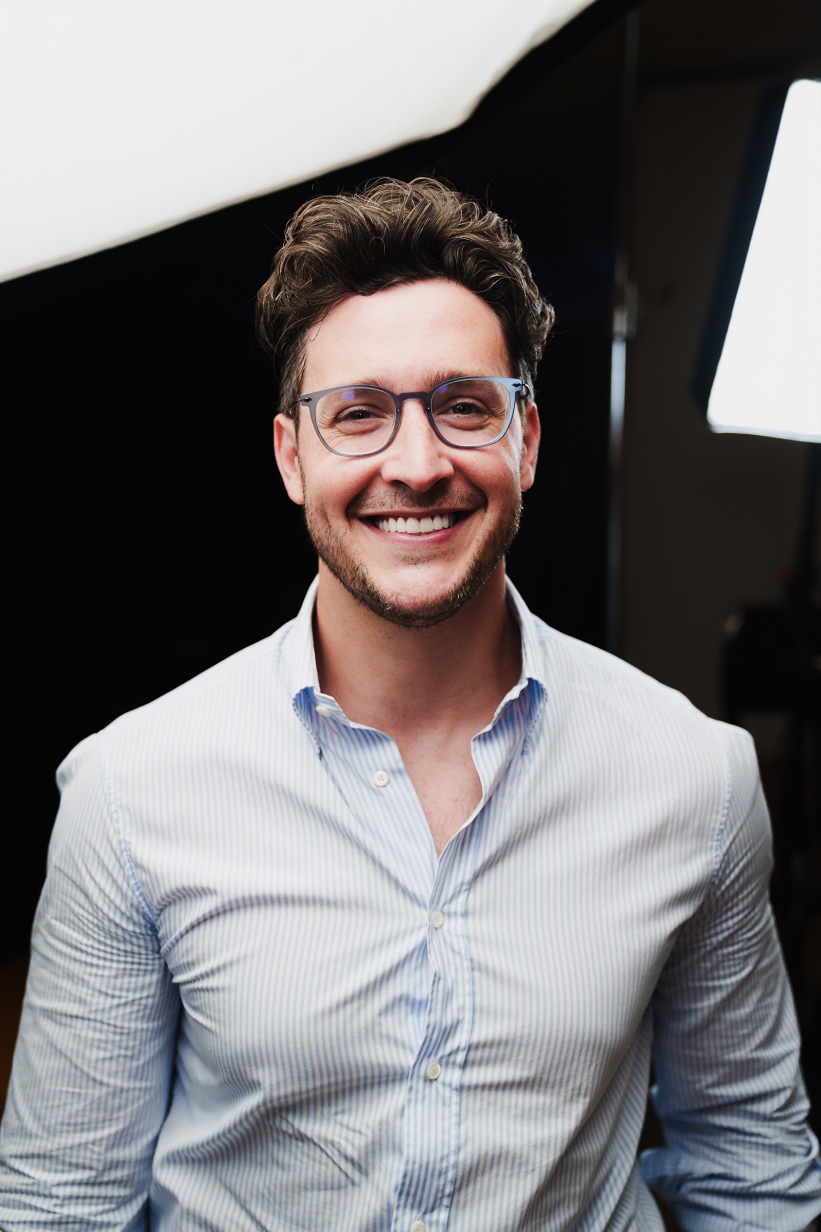 A man in a pale blue shirt and glasses smiles at the camera beside a softbox