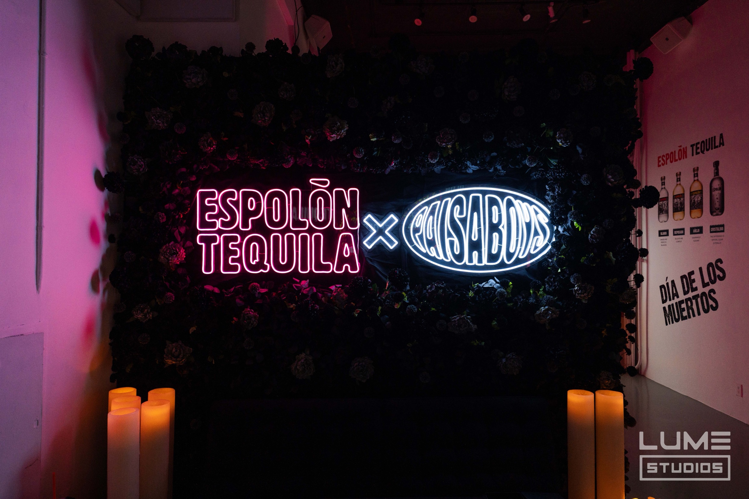 Pink neon Espolon Tequila sign beside a white neon oval logo on a wall of dark flowers above candles