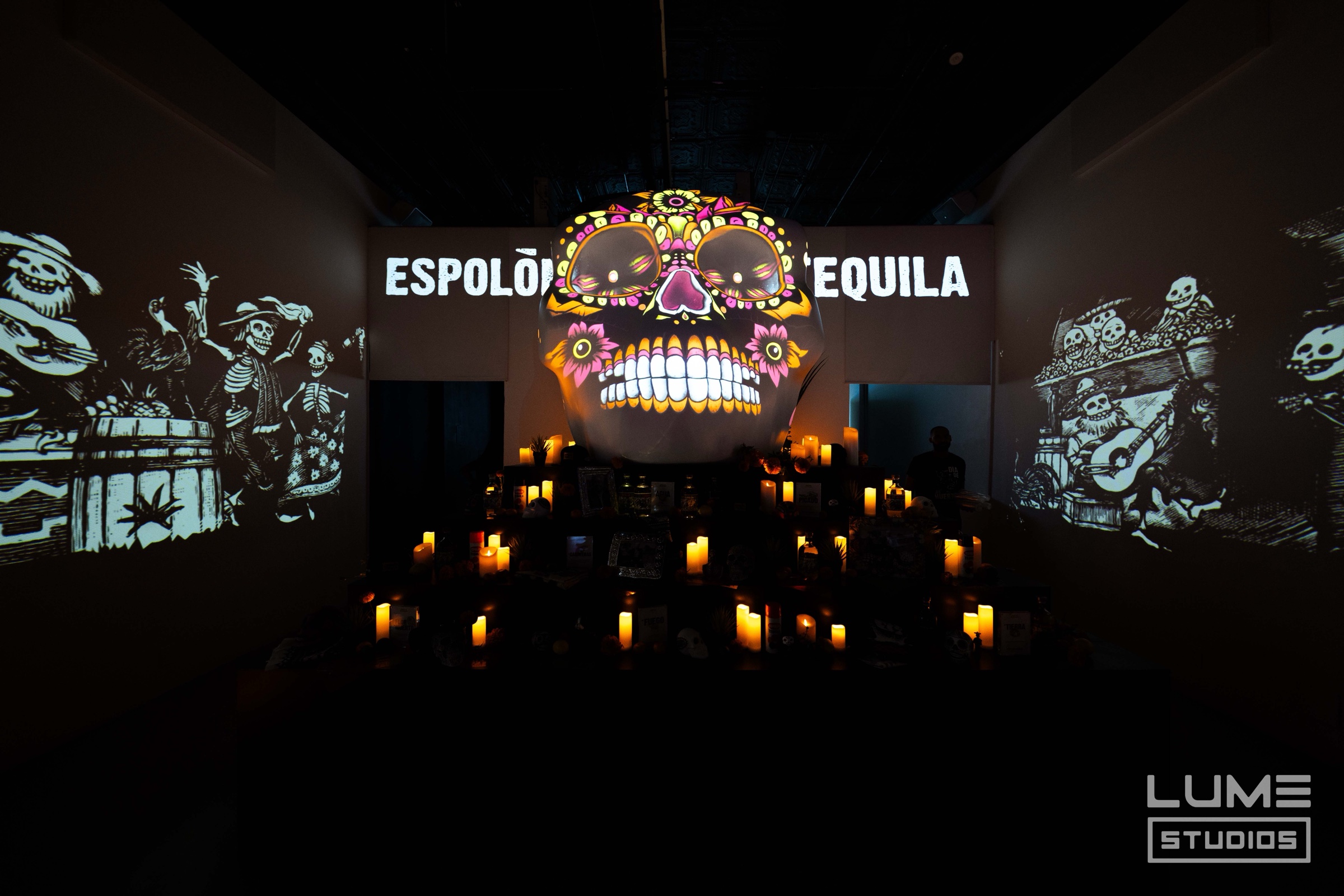 A colourful sugar skull and the words Espolon Tequila projected on a wall above rows of flickering candles