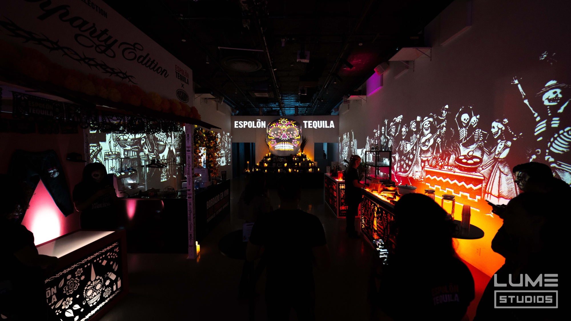 A dark hall with skeleton figures projected on the walls and an Espolon Tequila sign above a glowing skull