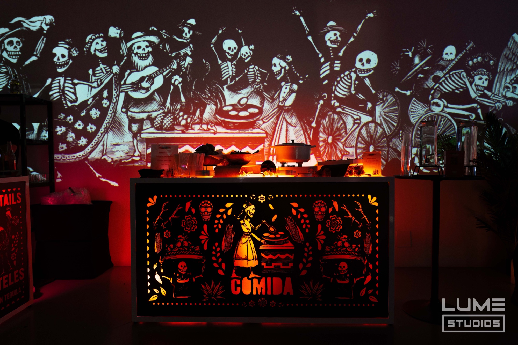 A glowing orange bar front cut with a dancing figure and the word Comida stands beneath projected skeleton musicians