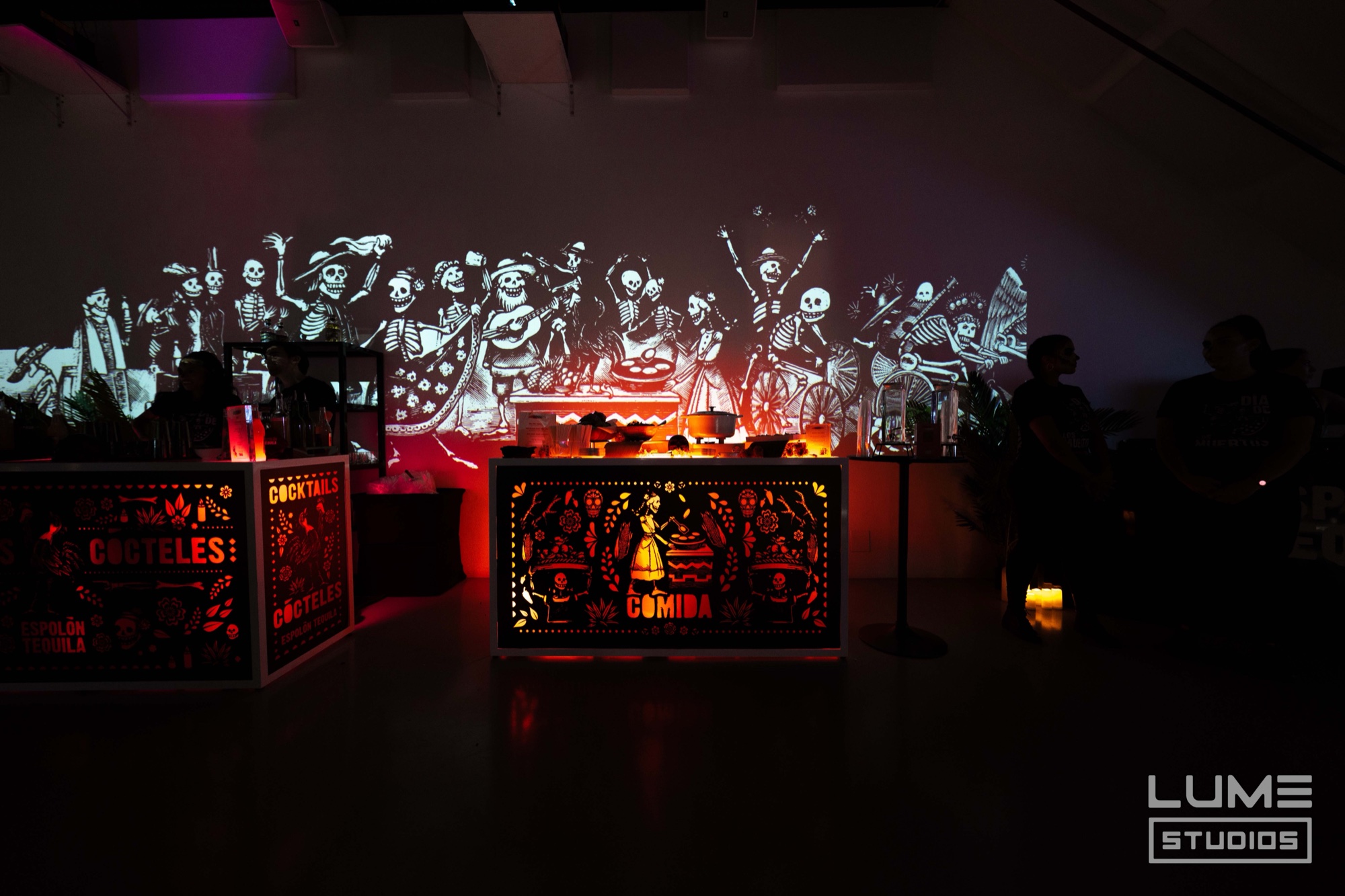 Illuminated papel picado style bars marked Cocteles and Comida below a projection of dancing skeleton figures