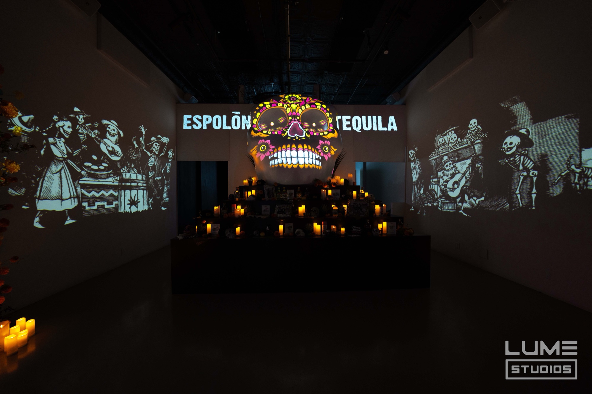 A glowing decorated sugar skull sits on tiers of candles beneath an Espolon Tequila sign, skeletons projected on both walls