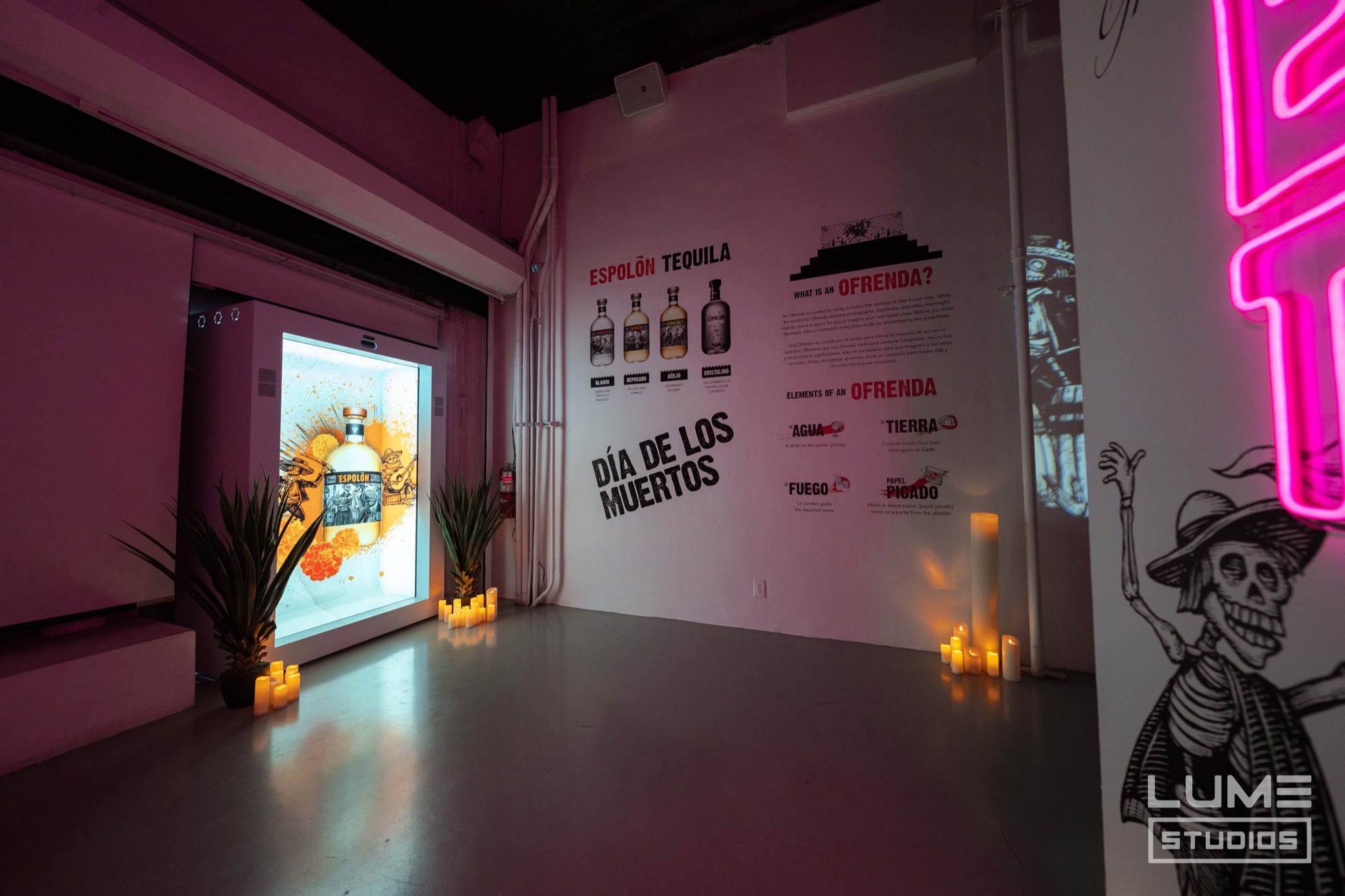 Lit Espolon bottles beside a pink wall printed with Dia de los Muertos text and a skeleton mural