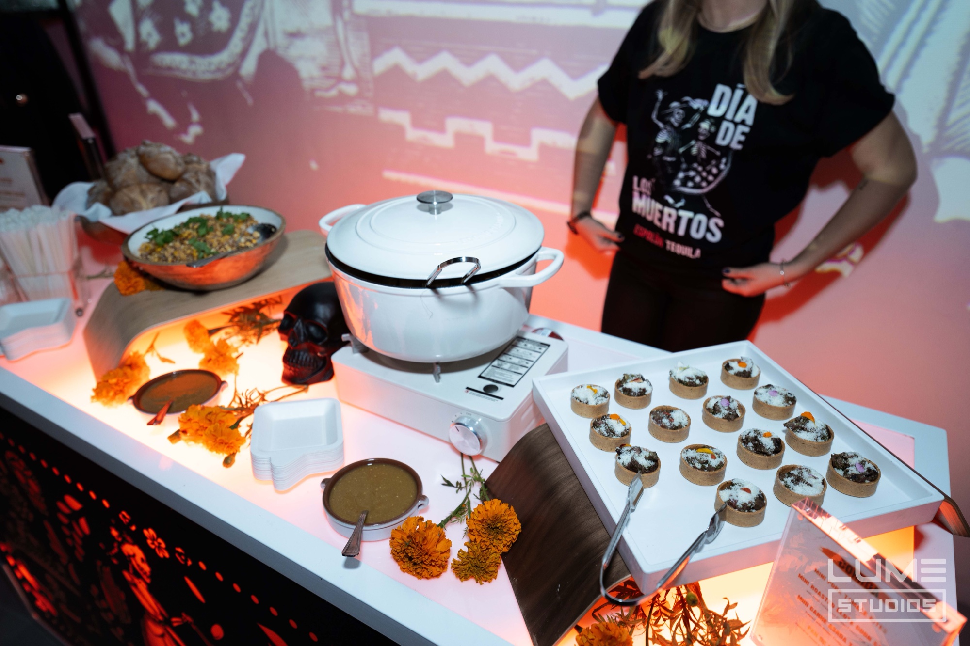 A server in a Dia de los Muertos shirt stands behind a buffet of tarts and marigolds under orange light