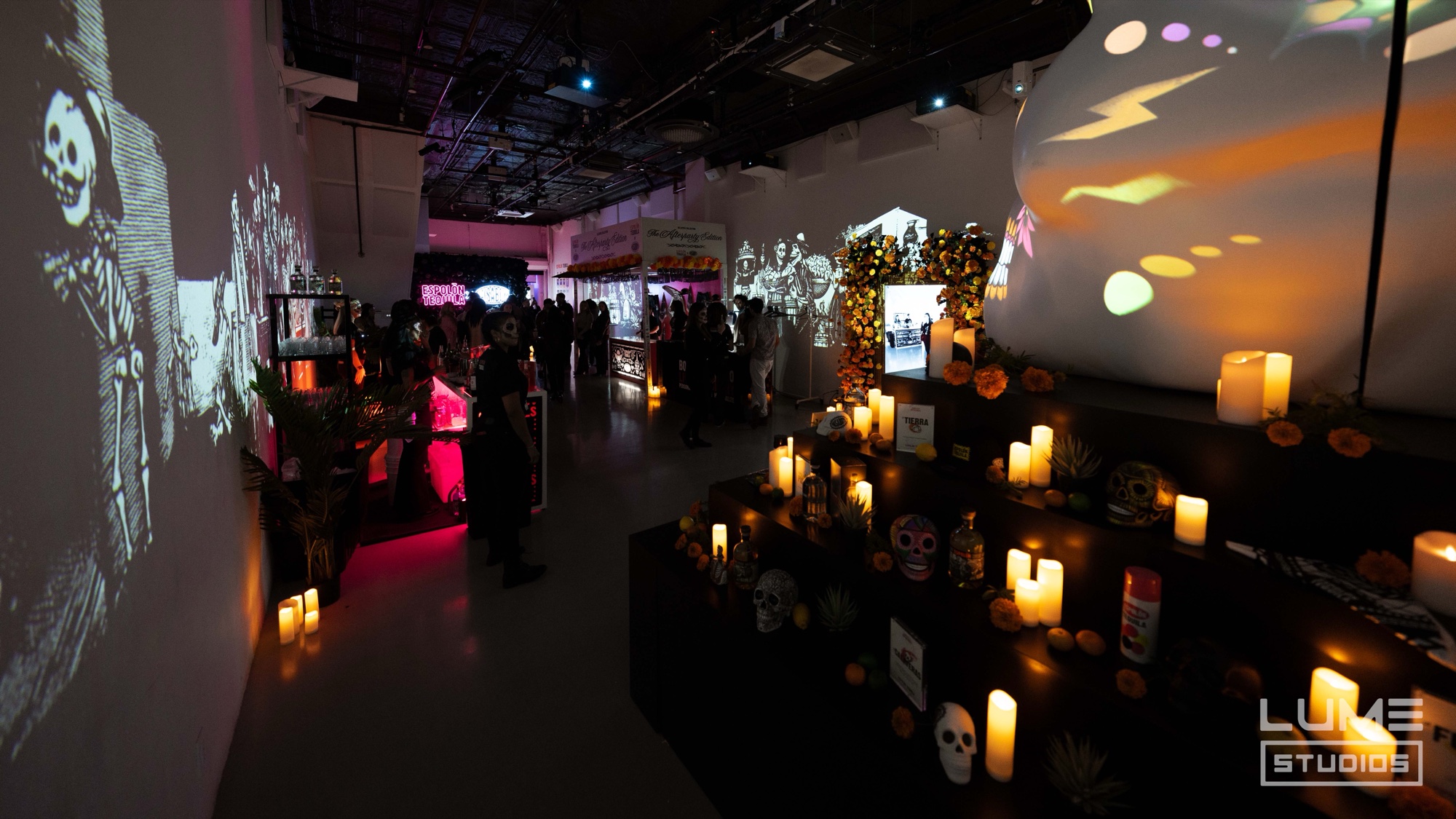 Guests wander past a dark altar of candles and sugar skulls with skeleton figures projected on the white walls
