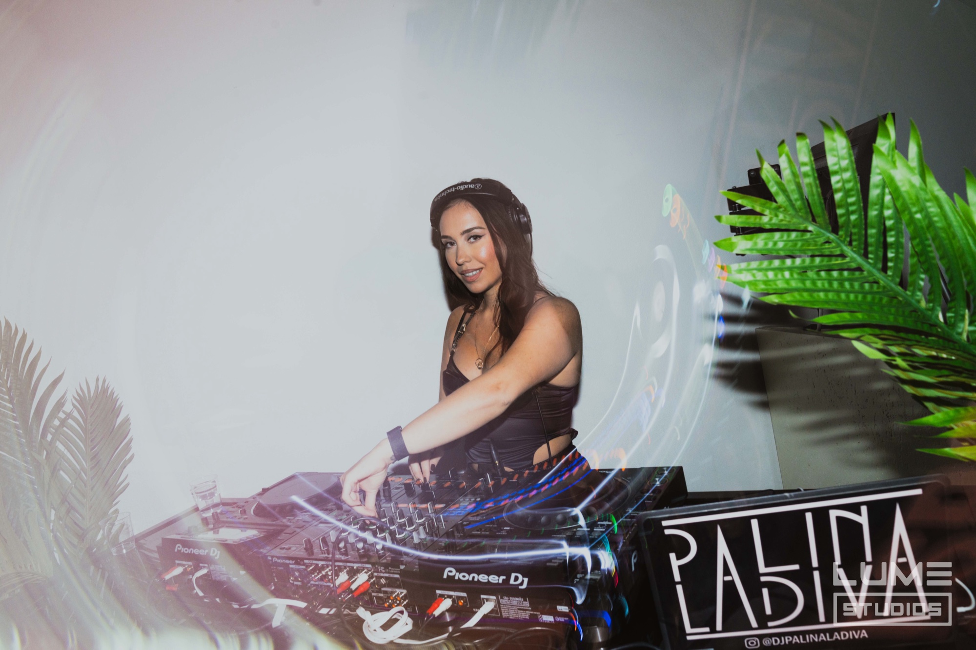 A DJ in a black top mixes on a Pioneer deck between palm fronds with a projected pattern behind her