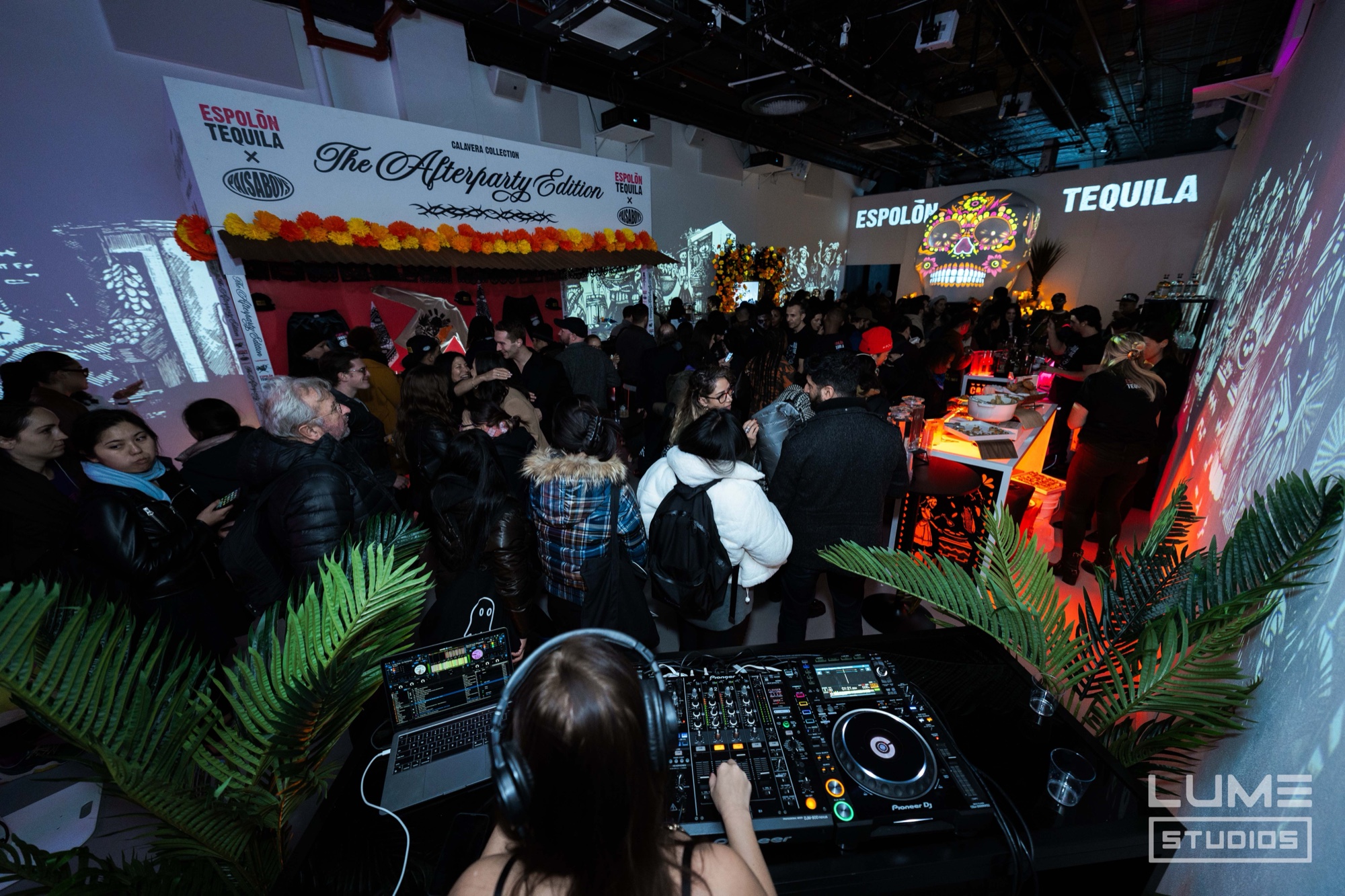 DJ in headphones at decks above a packed crowd beneath Espolon Tequila afterparty projections and palms