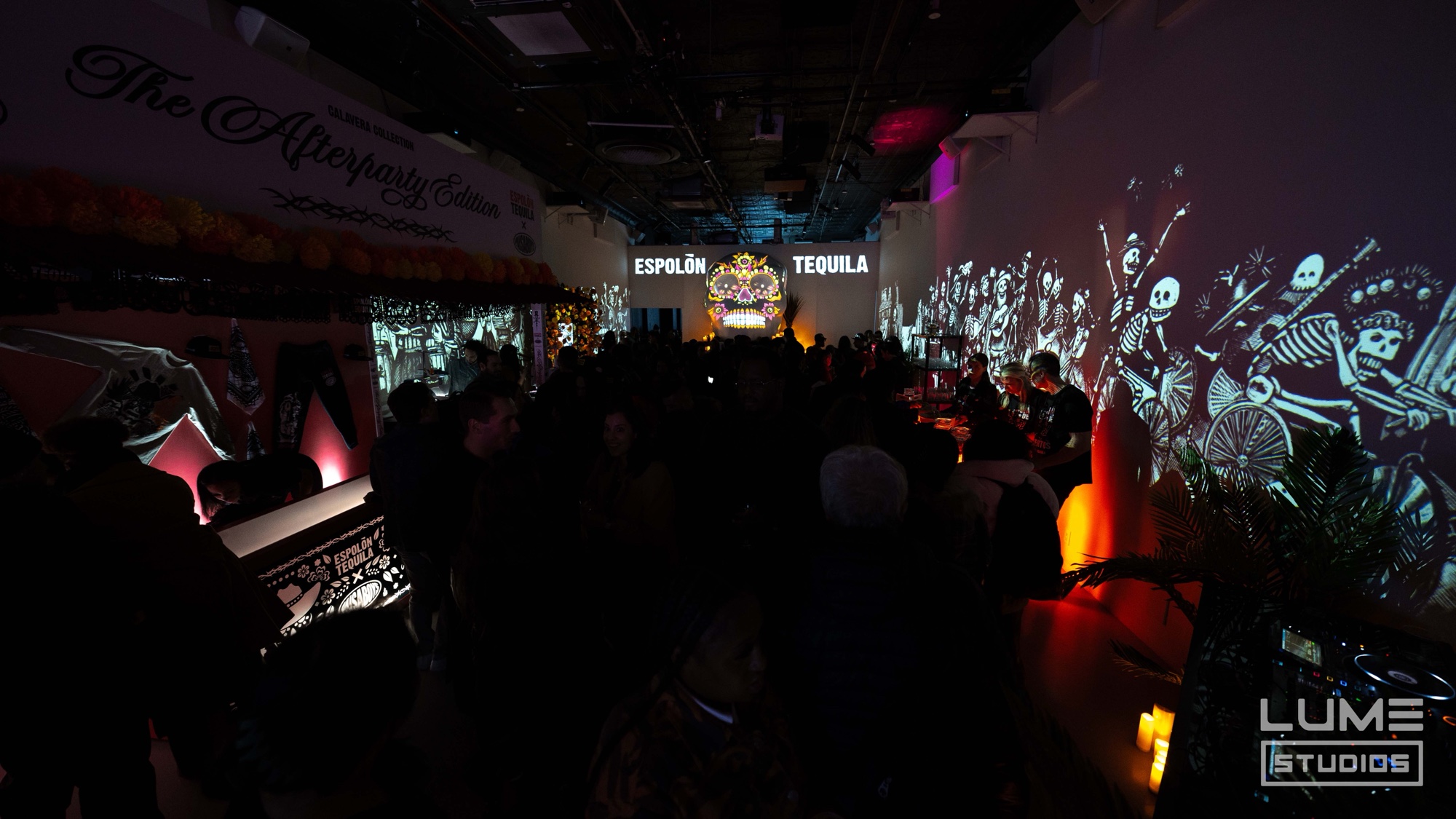 A crowd fills a dark venue lit by orange lamps with skeleton scenes and Espolon Tequila projected on the walls