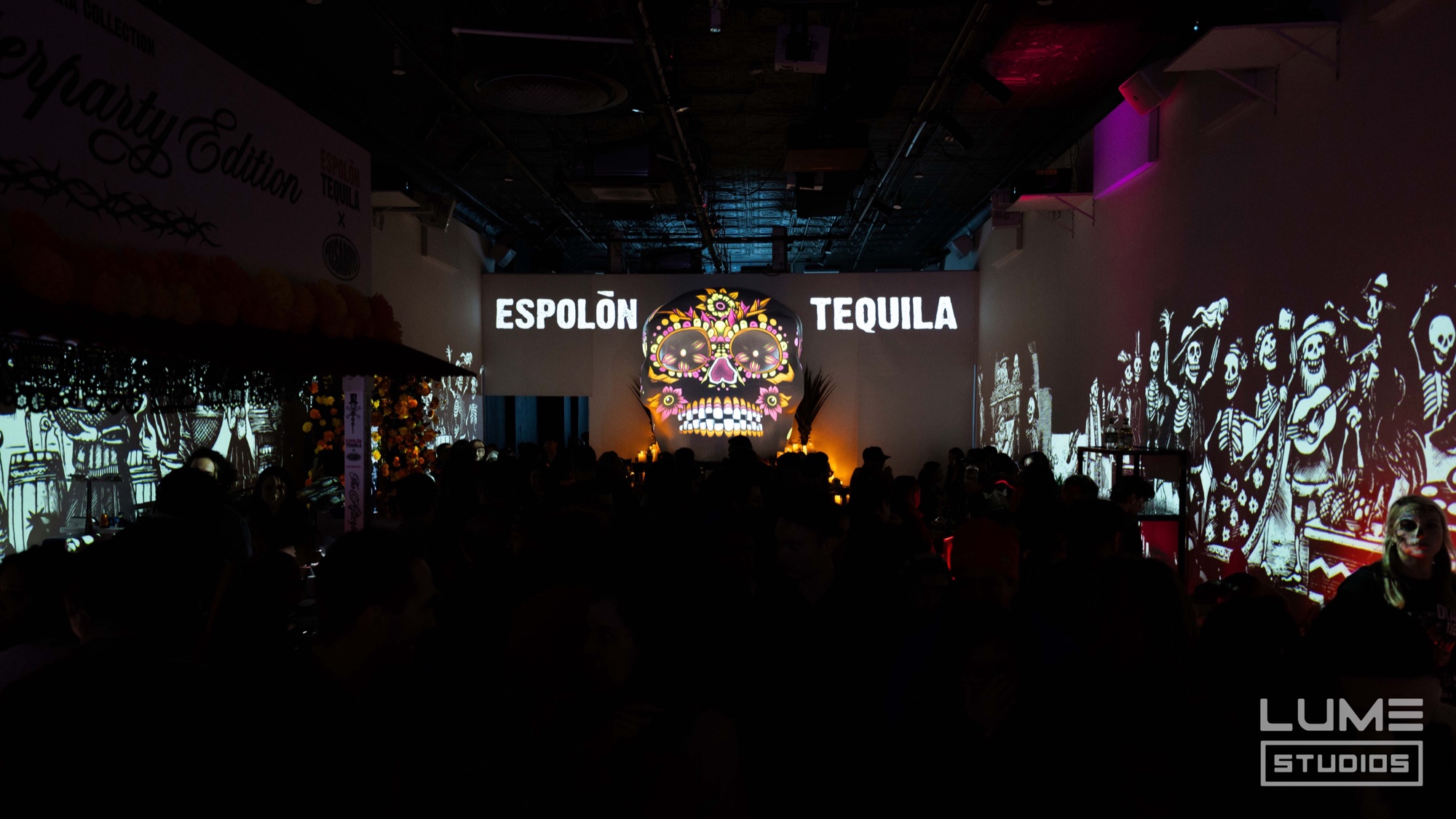 Silhouetted crowd faces a stage with an Espolon Tequila sign and lit skull display, skeleton murals on both walls