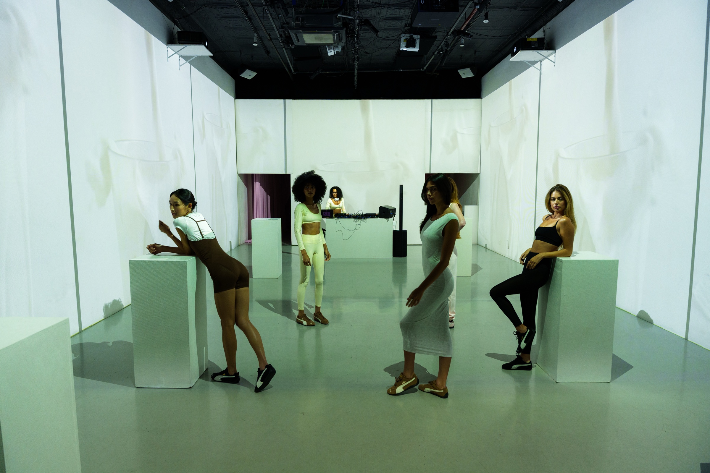 Four women in activewear pose on and beside pale green plinths in a white room with a DJ behind
