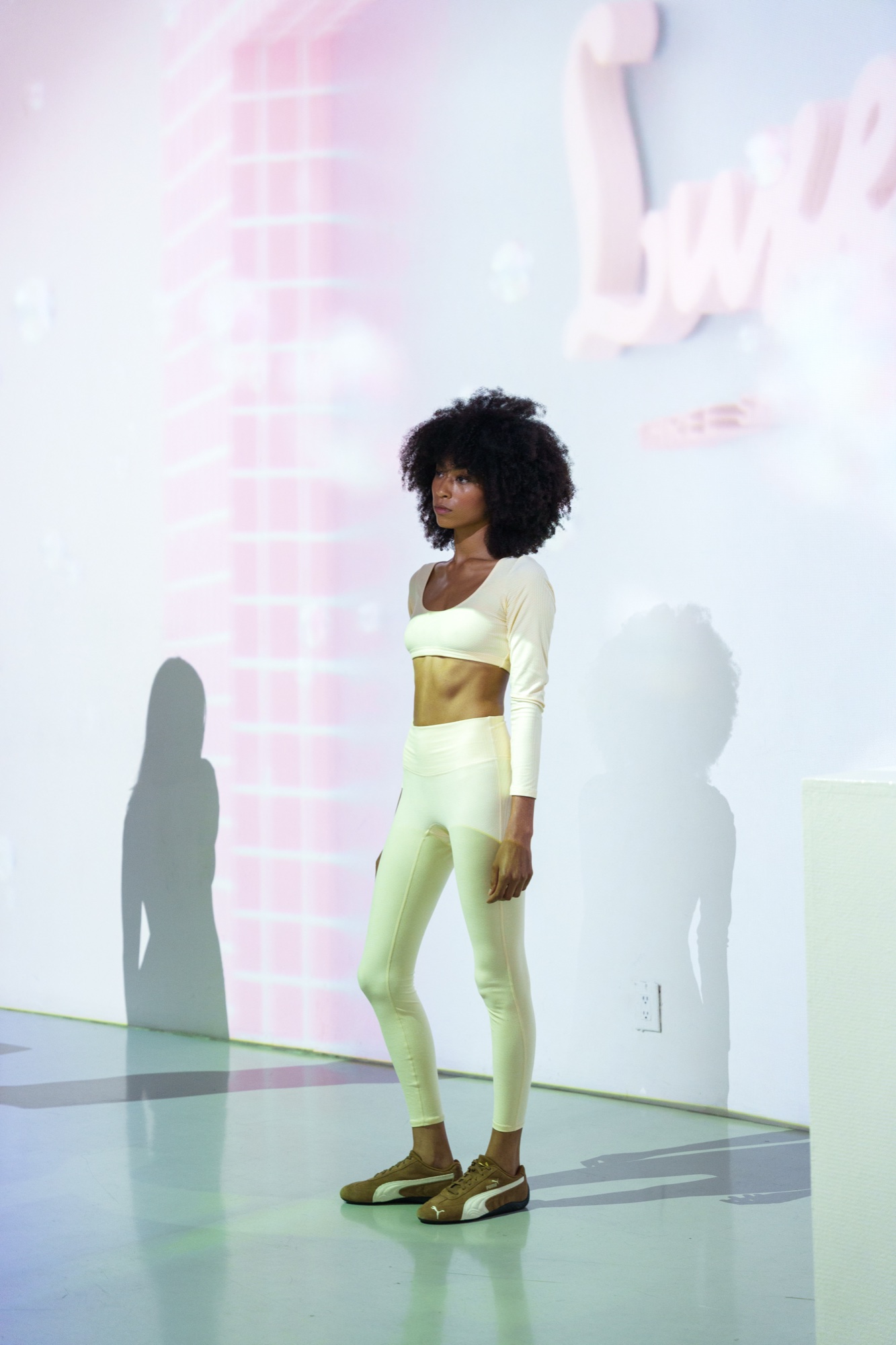 Model in a cream crop top and leggings stands before a pale pink projection with script lettering