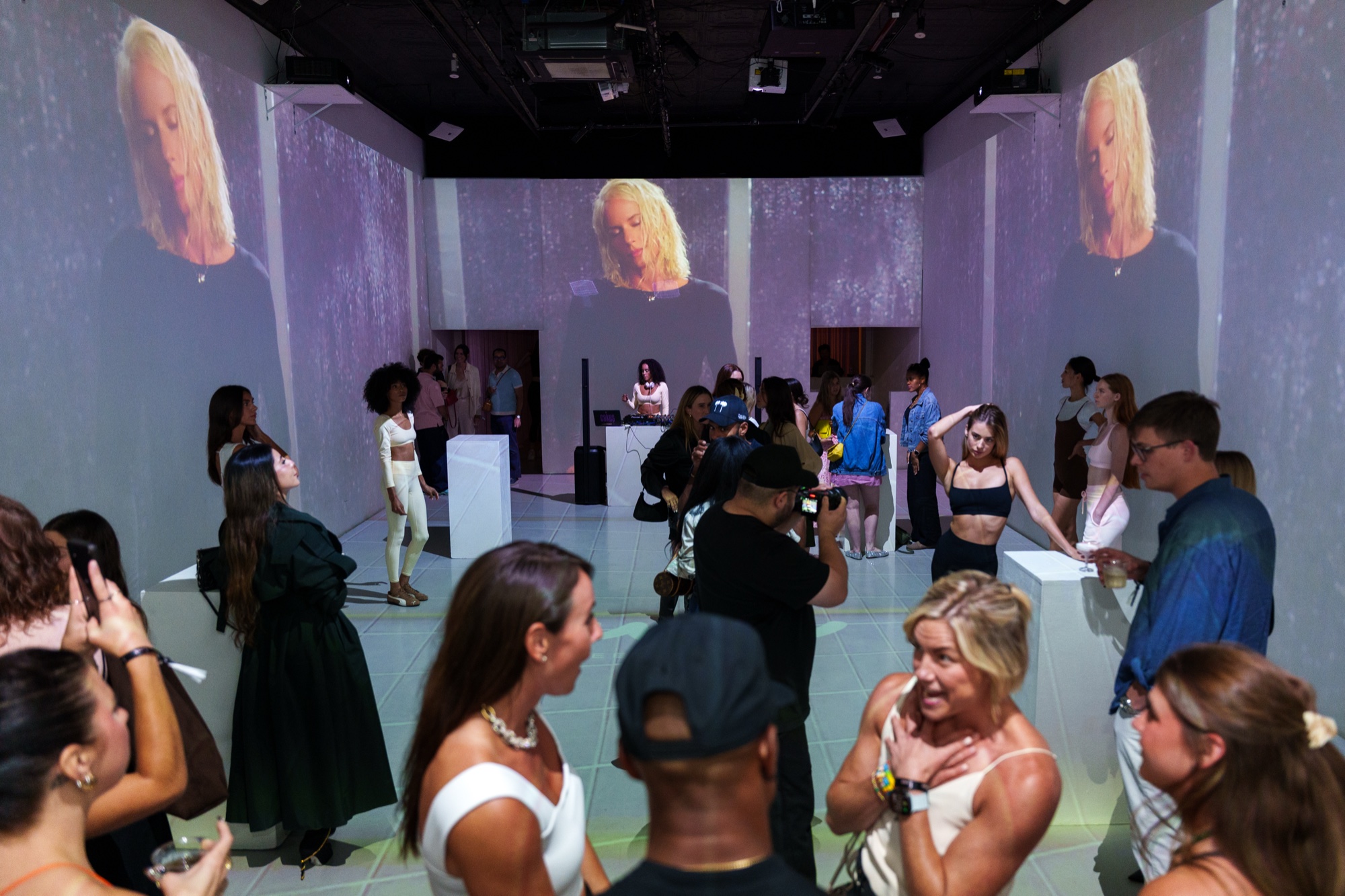Crowd of guests in a white room with a projected close up face on the far wall and DJ table