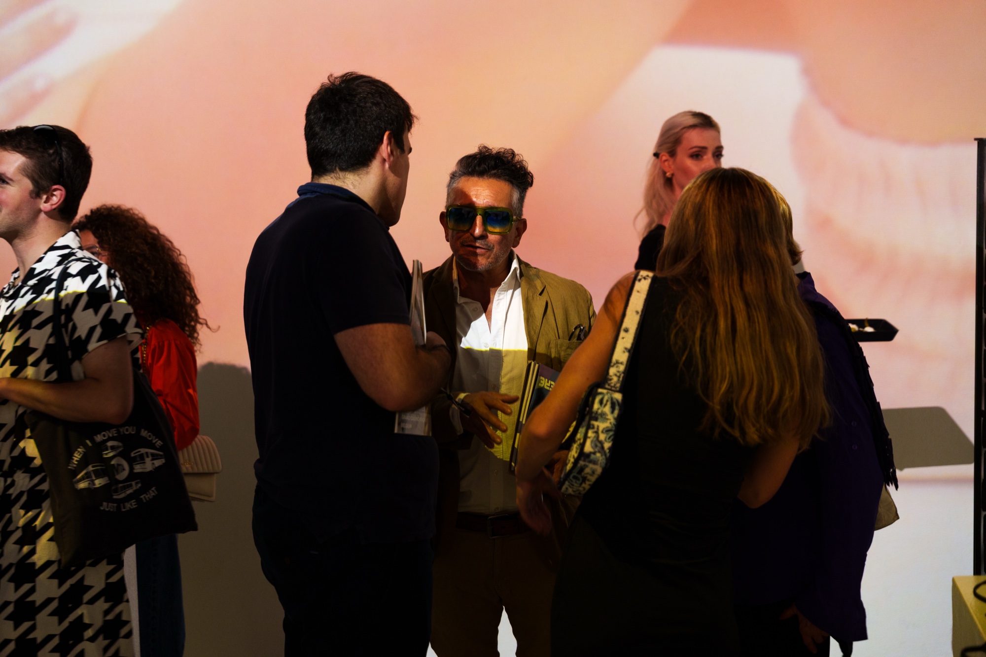 Man in a tan blazer and blue sunglasses chats with guests in front of a peach coloured projection