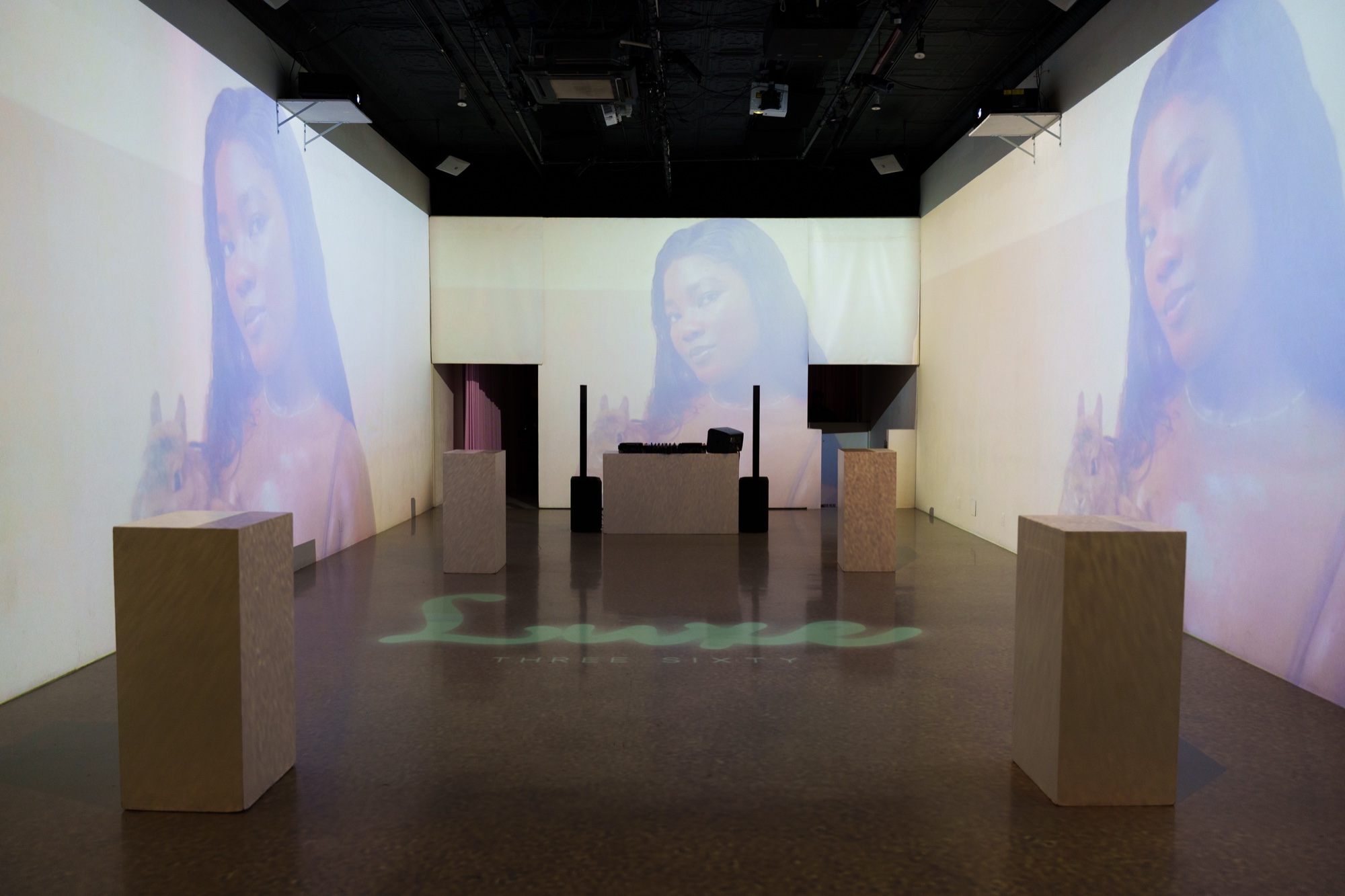Empty room with wooden plinths and a DJ booth surrounded by projected portraits of a woman with long dark hair