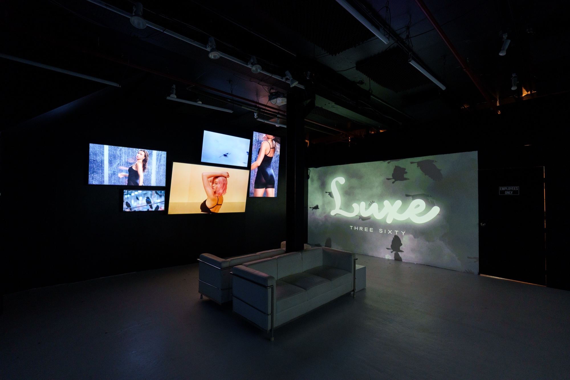 Dark room with a grey sofa facing screens of swimwear footage and a projected neon style Luxe logo