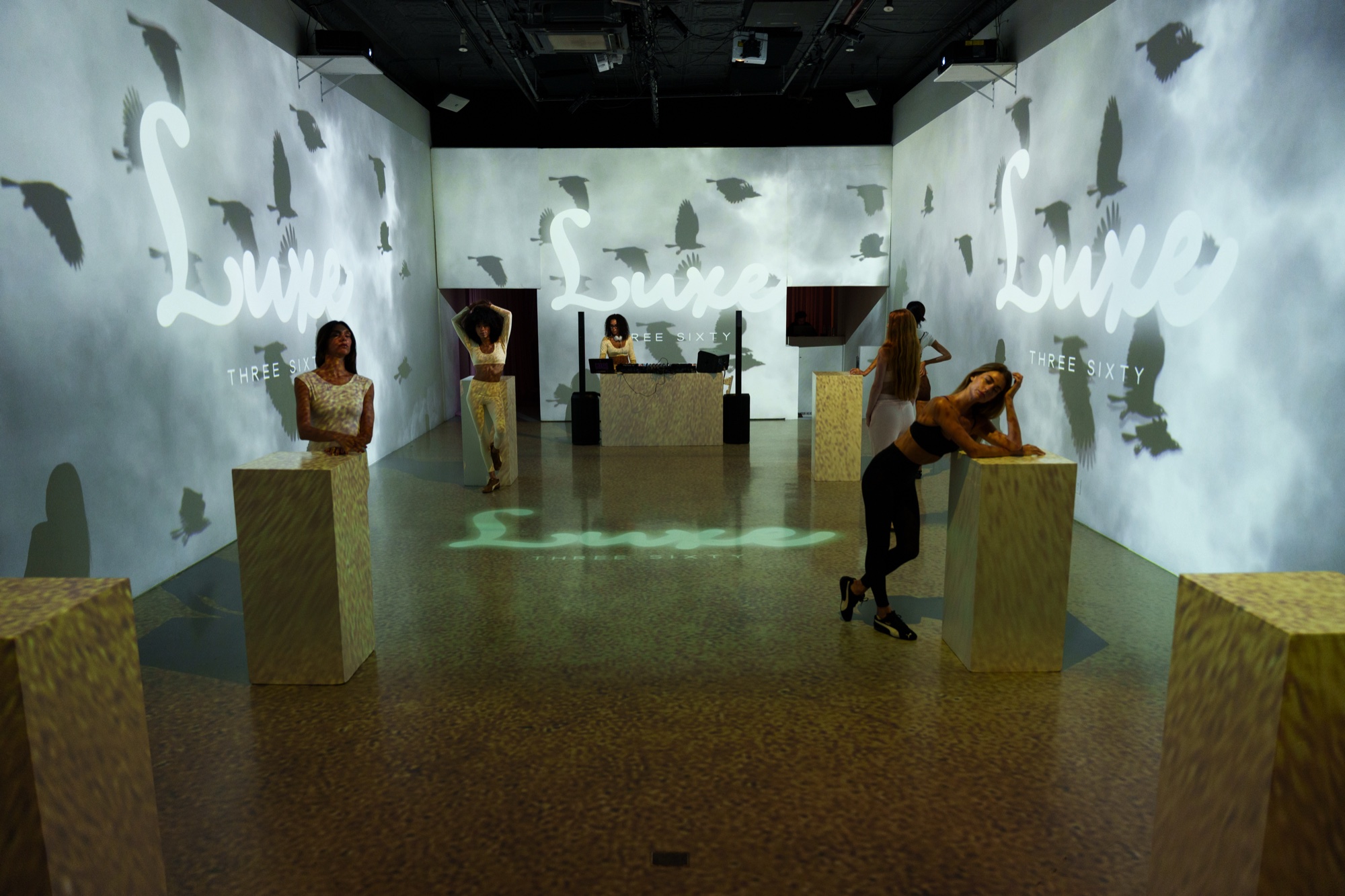 Models pose on cork plinths in a room projected with Luxe script lettering and drifting dark shapes