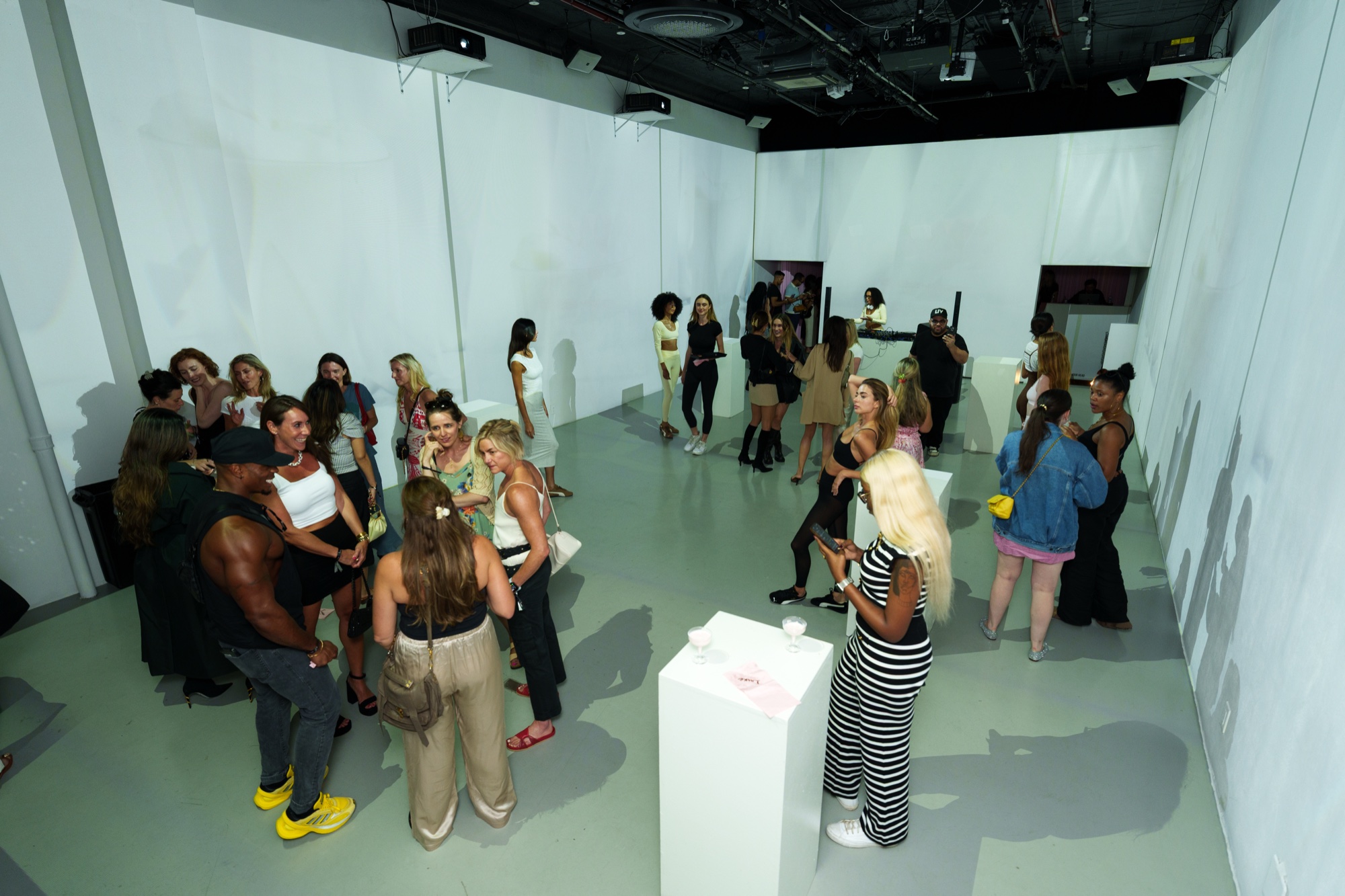 Crowd of guests mingles among white plinths in a bright white room seen from above