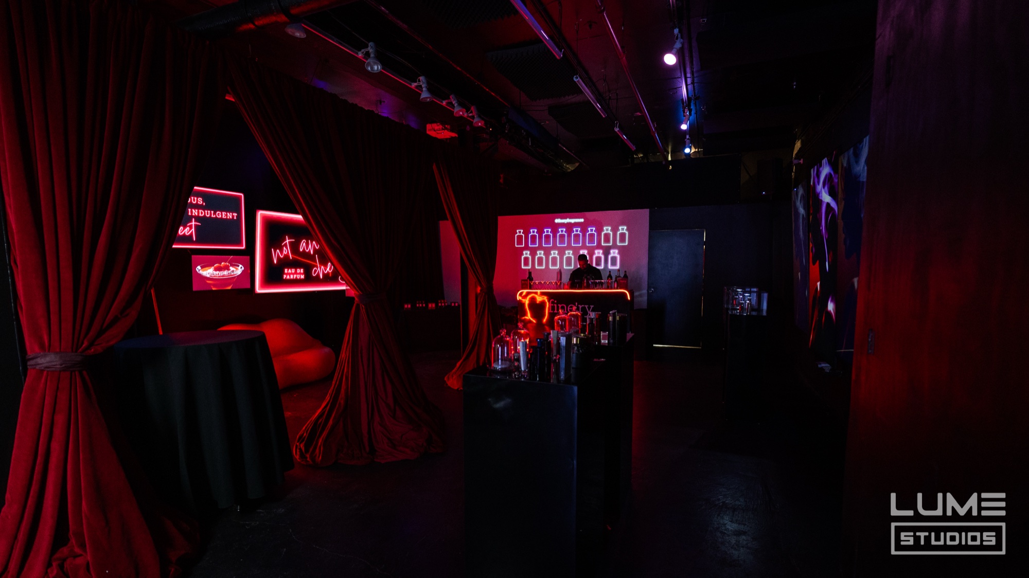 A dark lounge with red velvet curtains, a lit bar and pink neon signs glowing on a black wall
