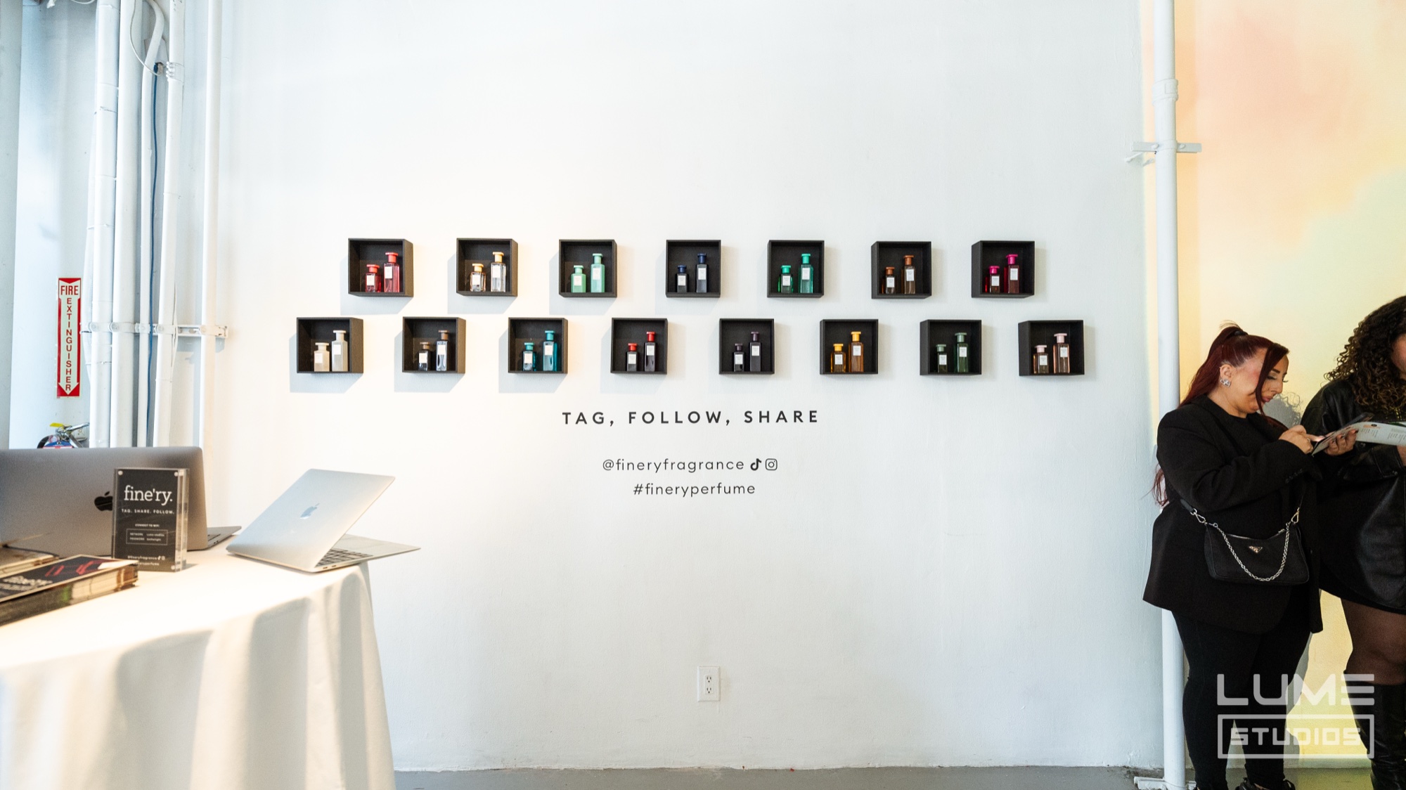Fourteen small black shadow boxes of perfume bottles on a white wall above the words Tag Follow Share