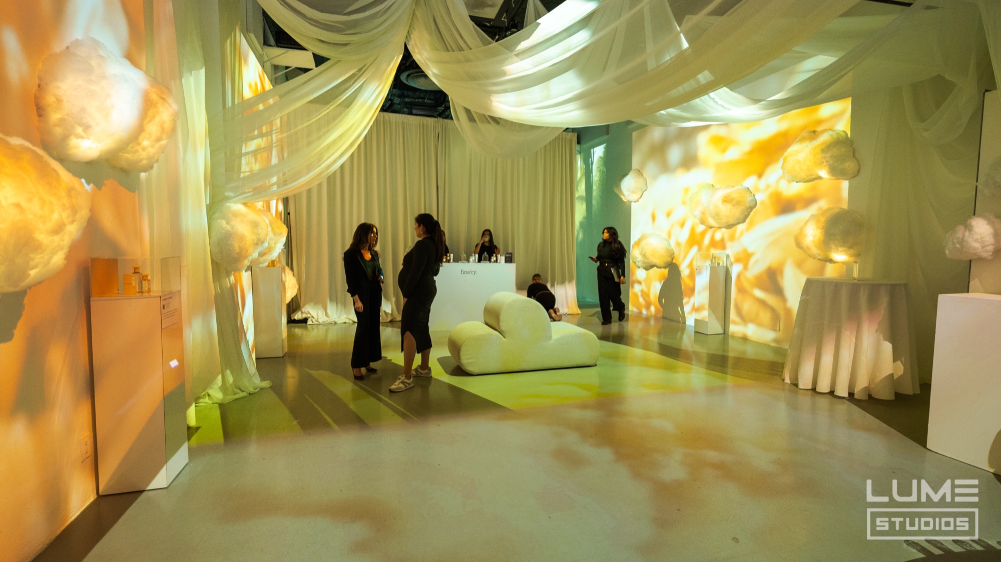Guests stand among white pedestals under draped white fabric while yellow lemon imagery is projected across the walls