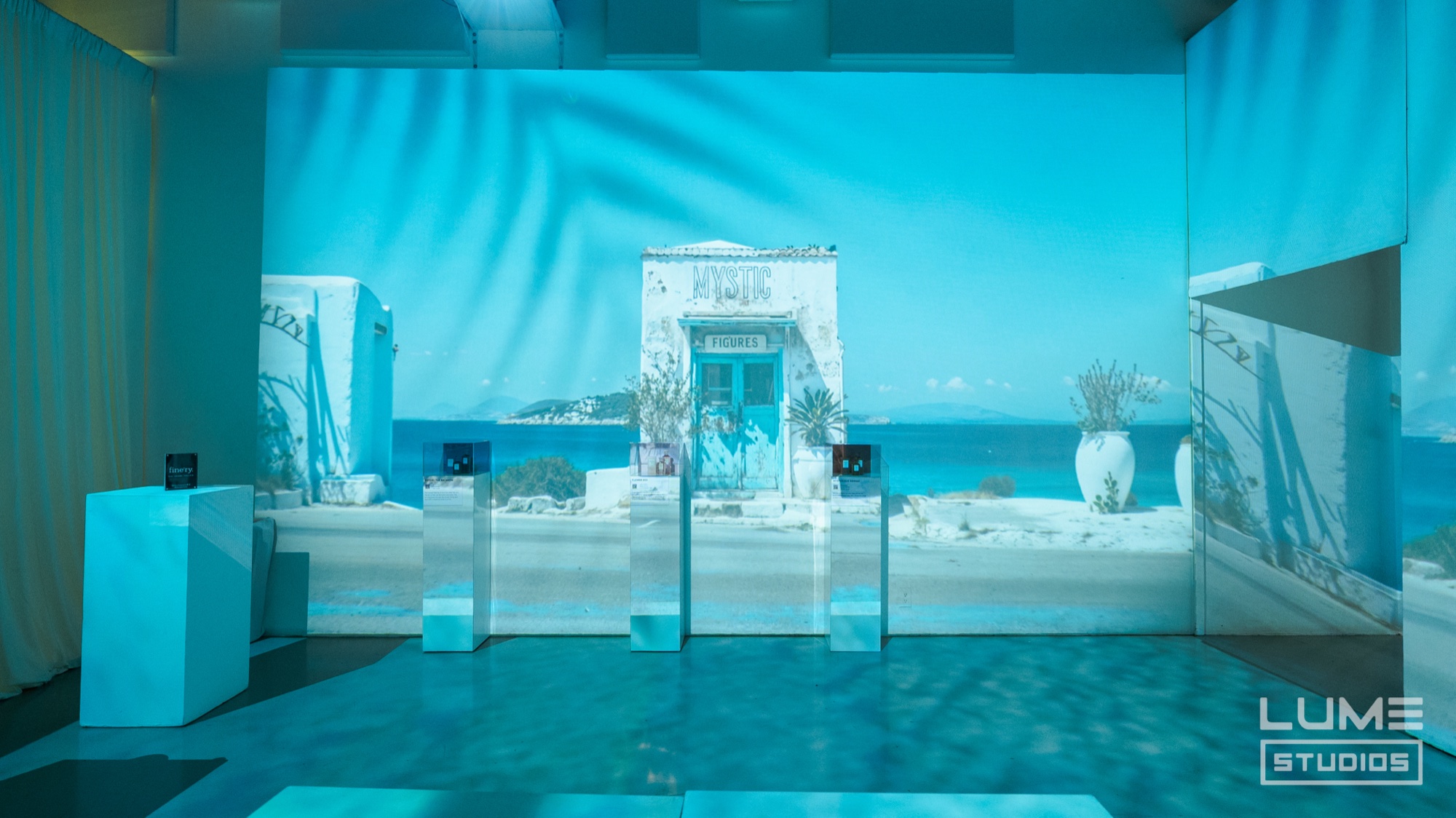 Blue toned room with mirrored plinths before a projection of a white Greek island doorway and sea