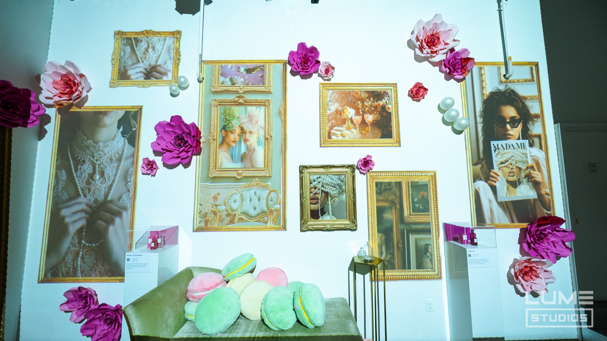 Gold framed portraits and pink paper flowers cover a white wall above a sofa piled with giant pastel macaron cushions