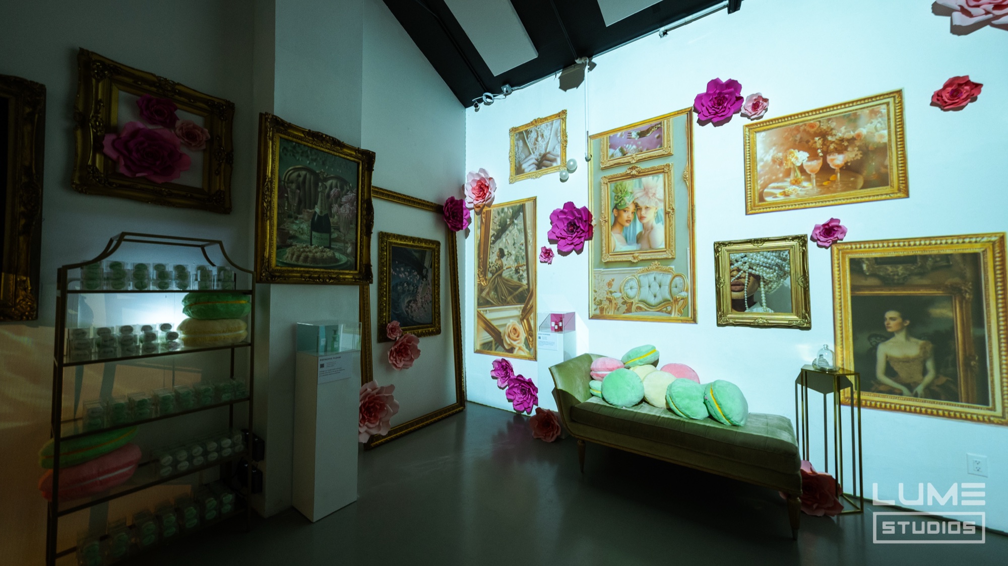 Gilt framed paintings and pink paper flowers cover a white wall above a bench of pastel cushions
