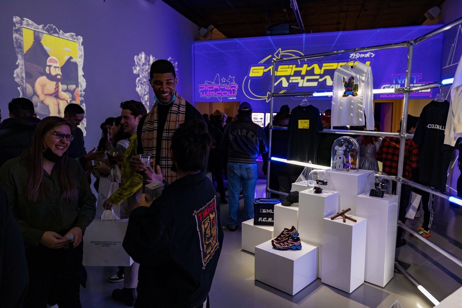 Guests chat beside white pedestals holding sneakers and collectibles under violet Gashapon Arcade projections