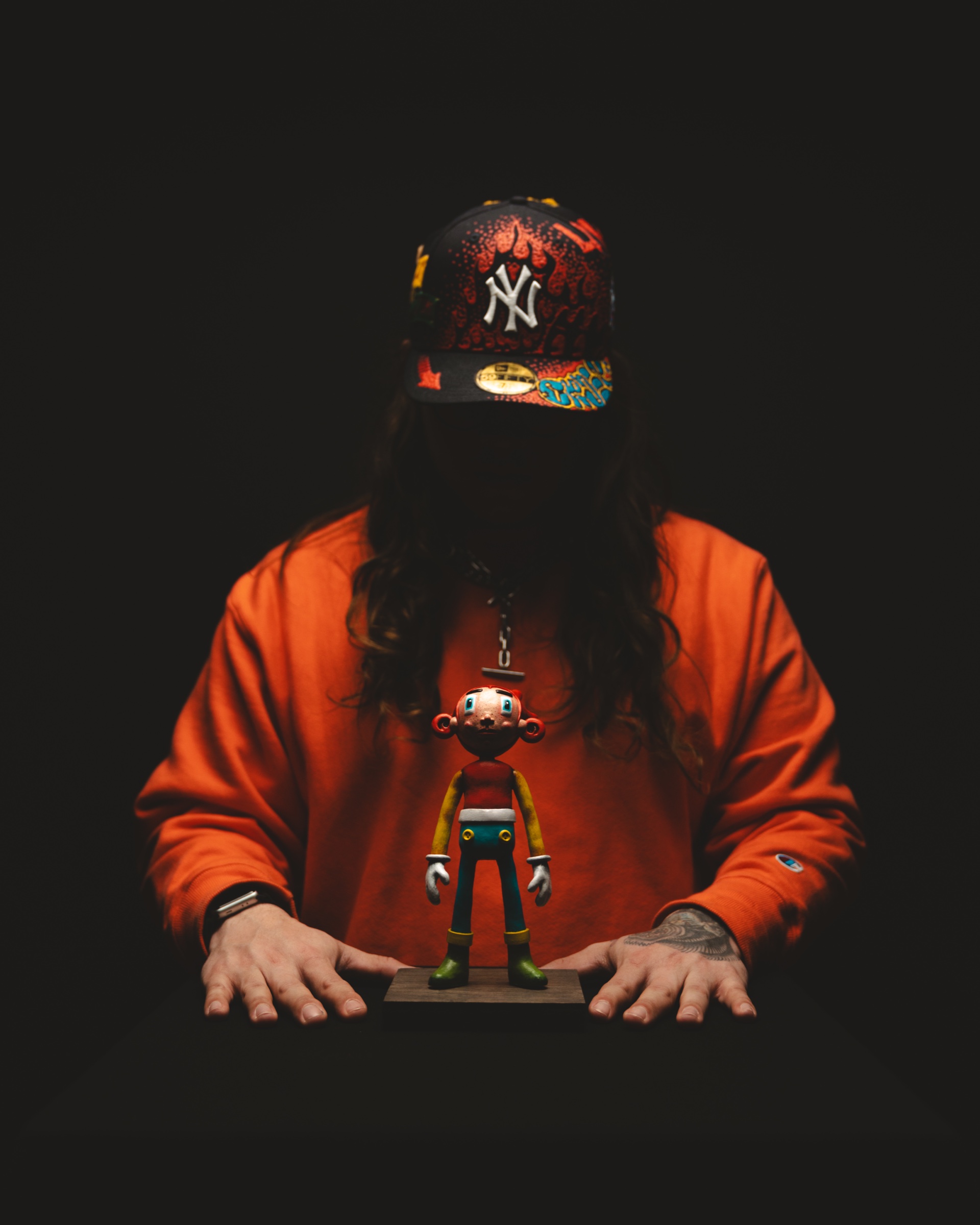 Pedro J Baez in an orange sweatshirt and New York cap behind a small Atomo figurine on a plinth