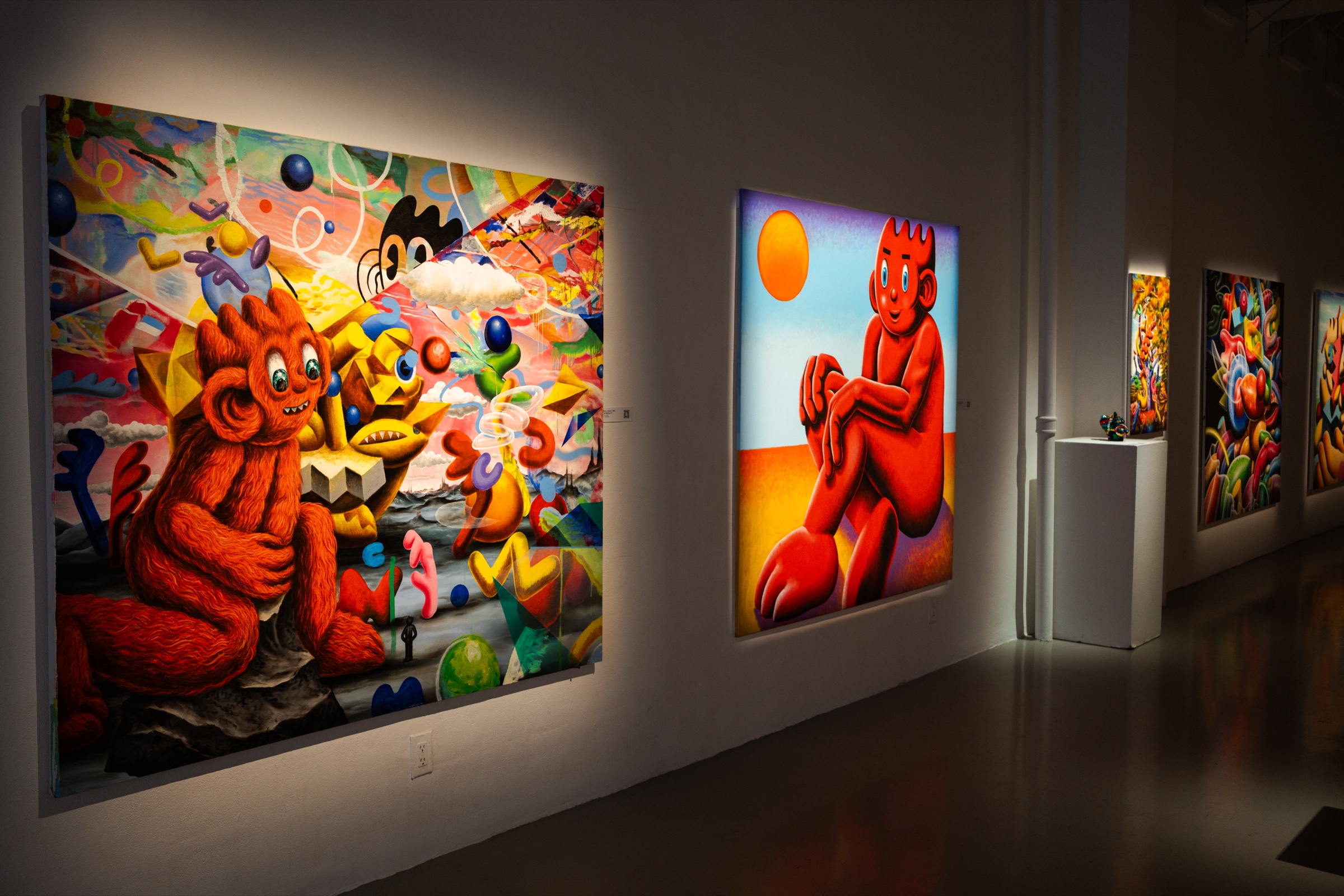 Orange creature paintings line a lit wall with a white plinth holding a sculpture further down the room