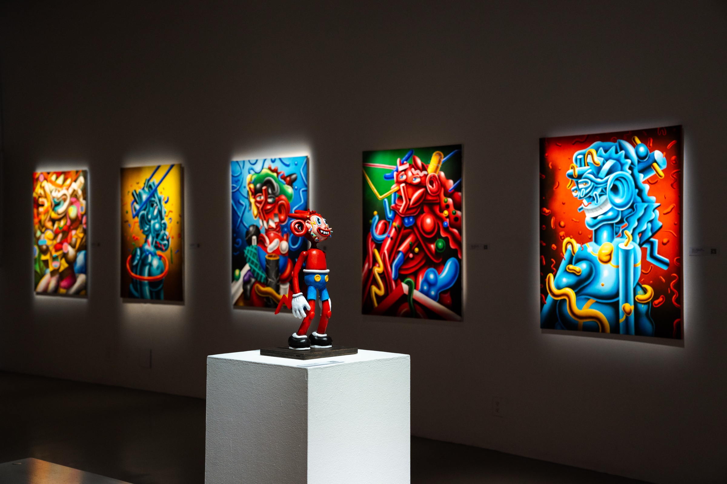 Five backlit paintings on a dark wall surround a red and blue Atomo sculpture on a white plinth