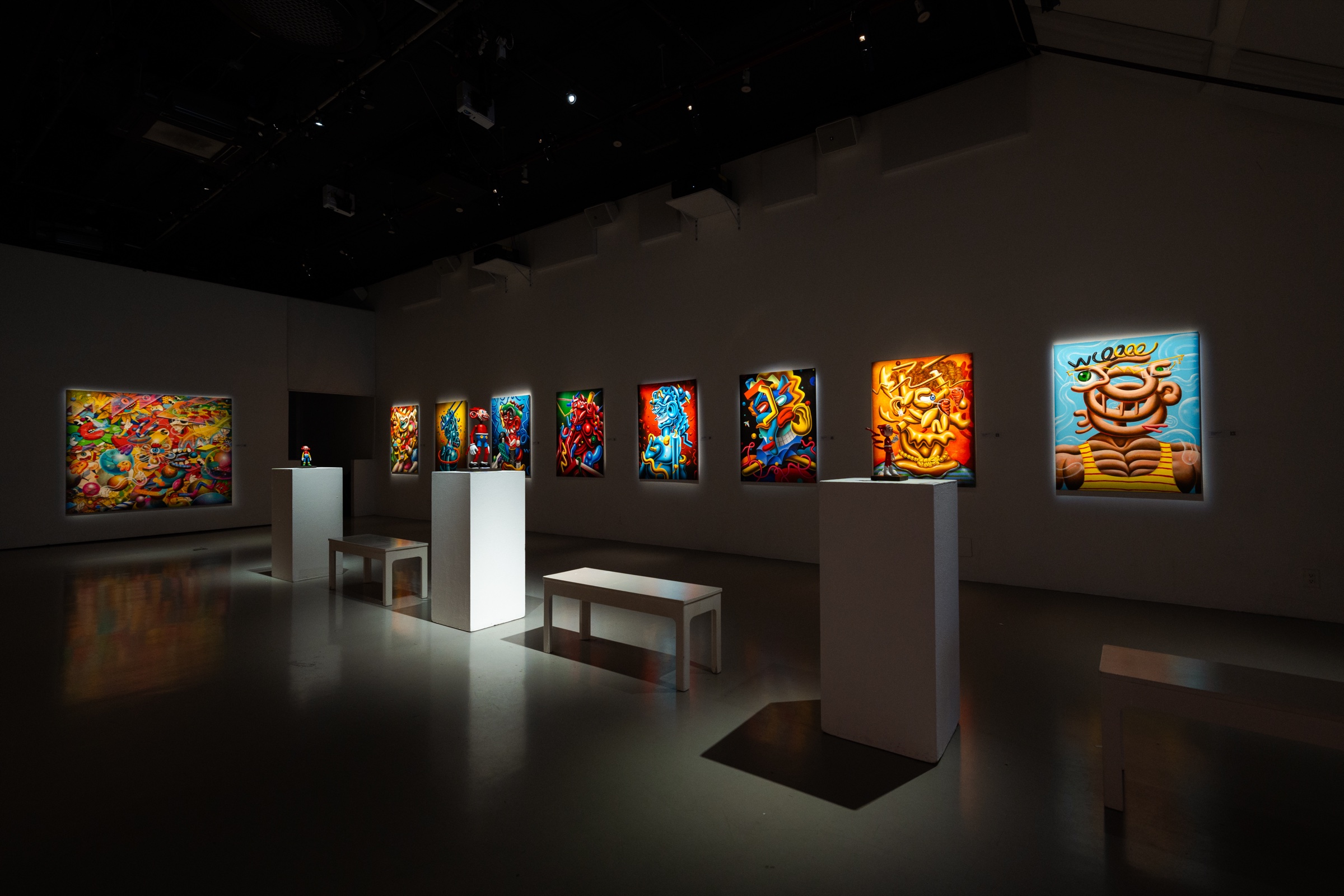 Wide gallery view of glowing canvases along two walls with plinths and a bench on a polished floor