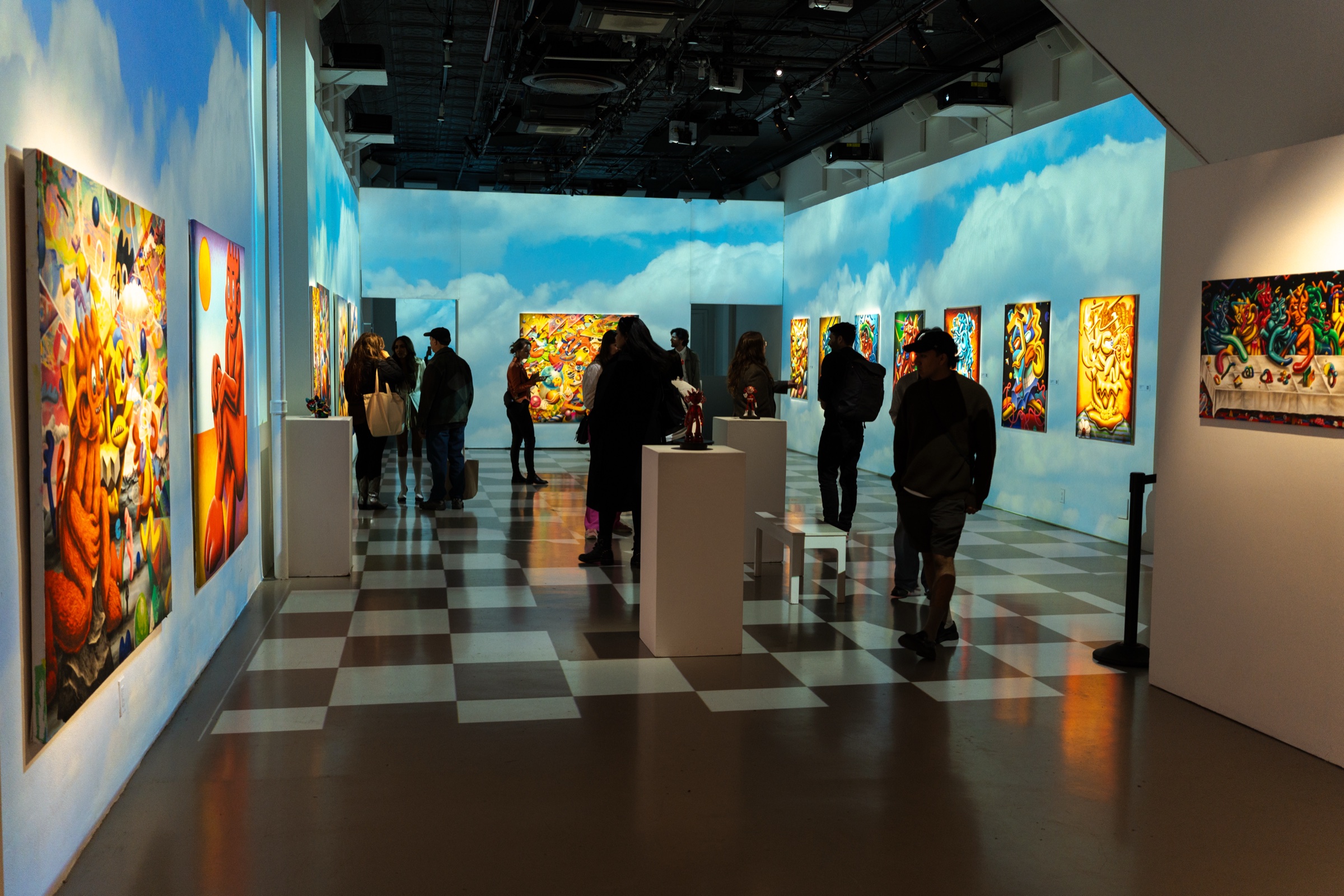 Visitors stand among canvases and plinths in a room washed with projected clouds above a checkered floor