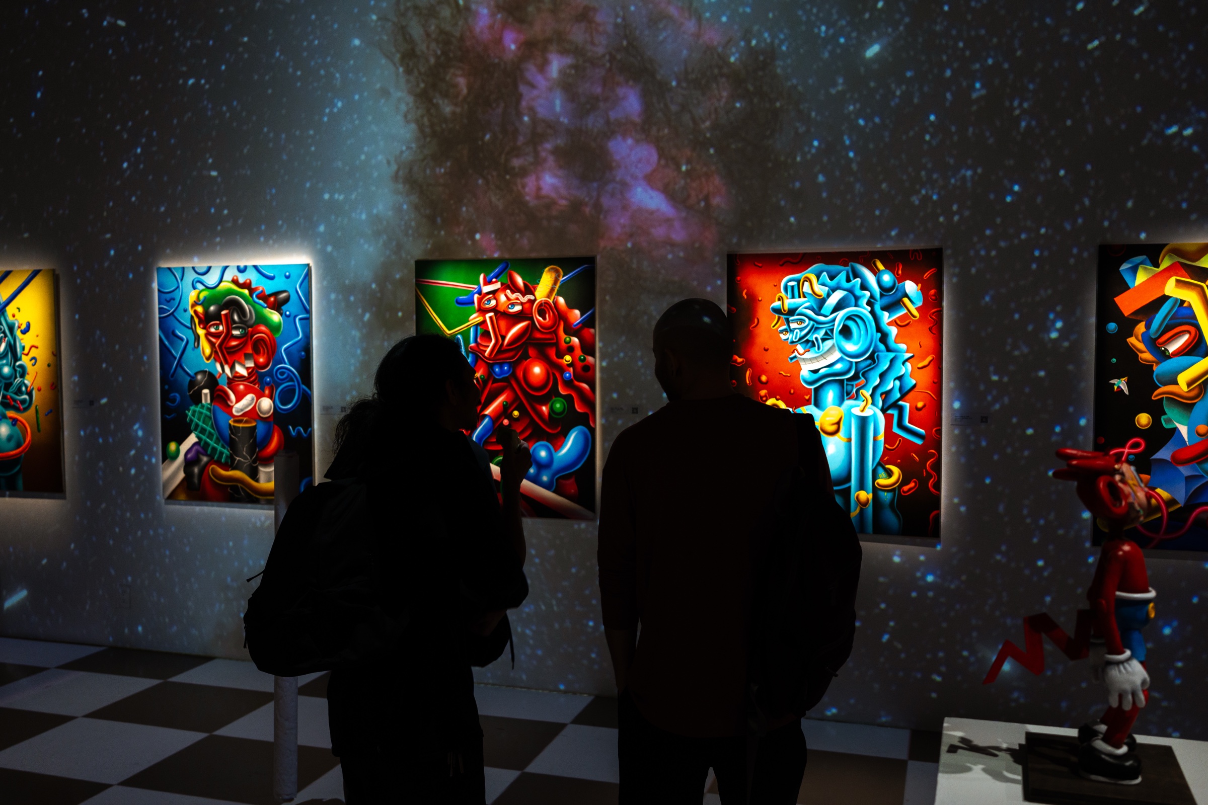 Two visitors in silhouette face backlit paintings in a room projected with stars and violet nebula