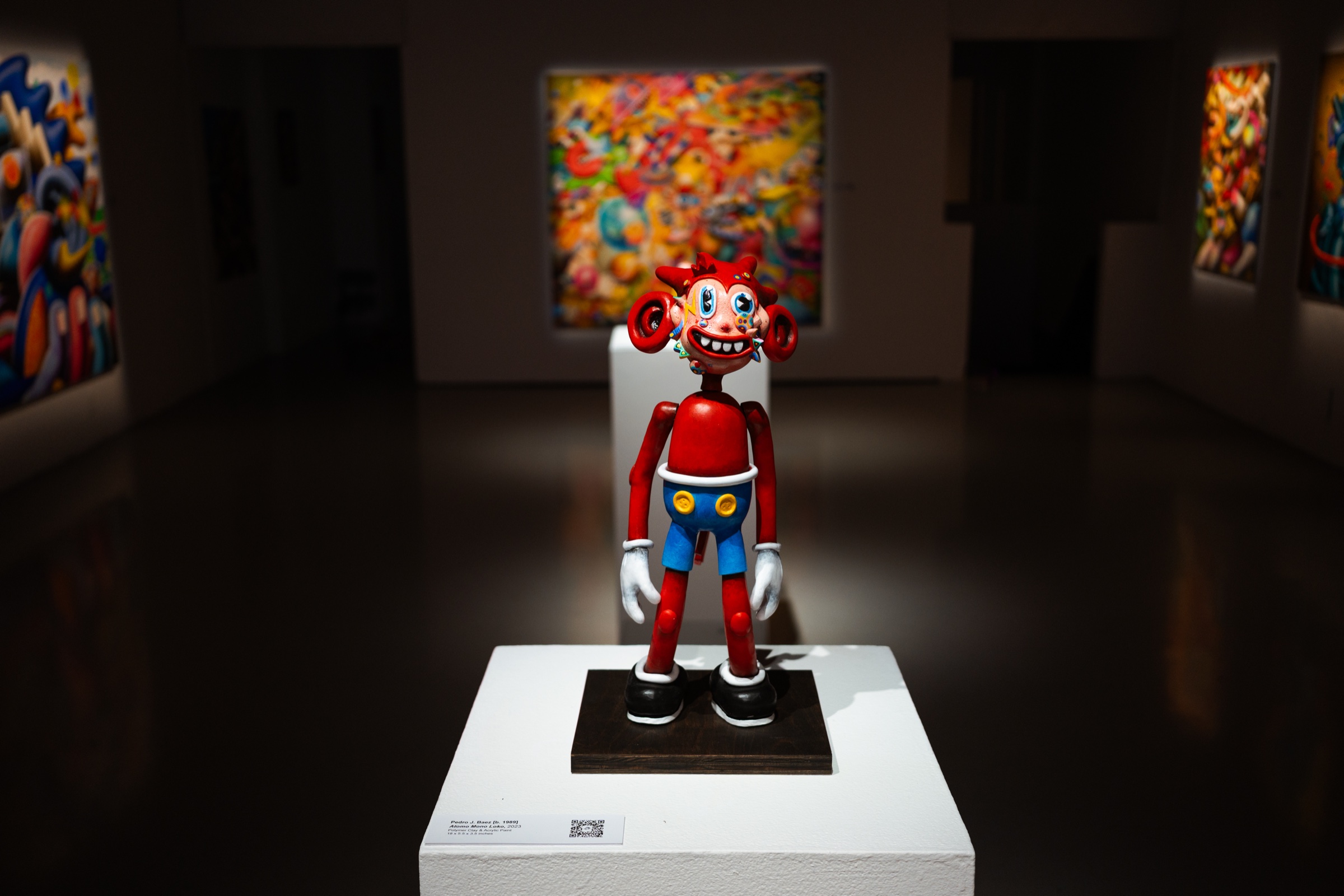A red and blue Atomo sculpture on a white plinth with a bright painting glowing behind it