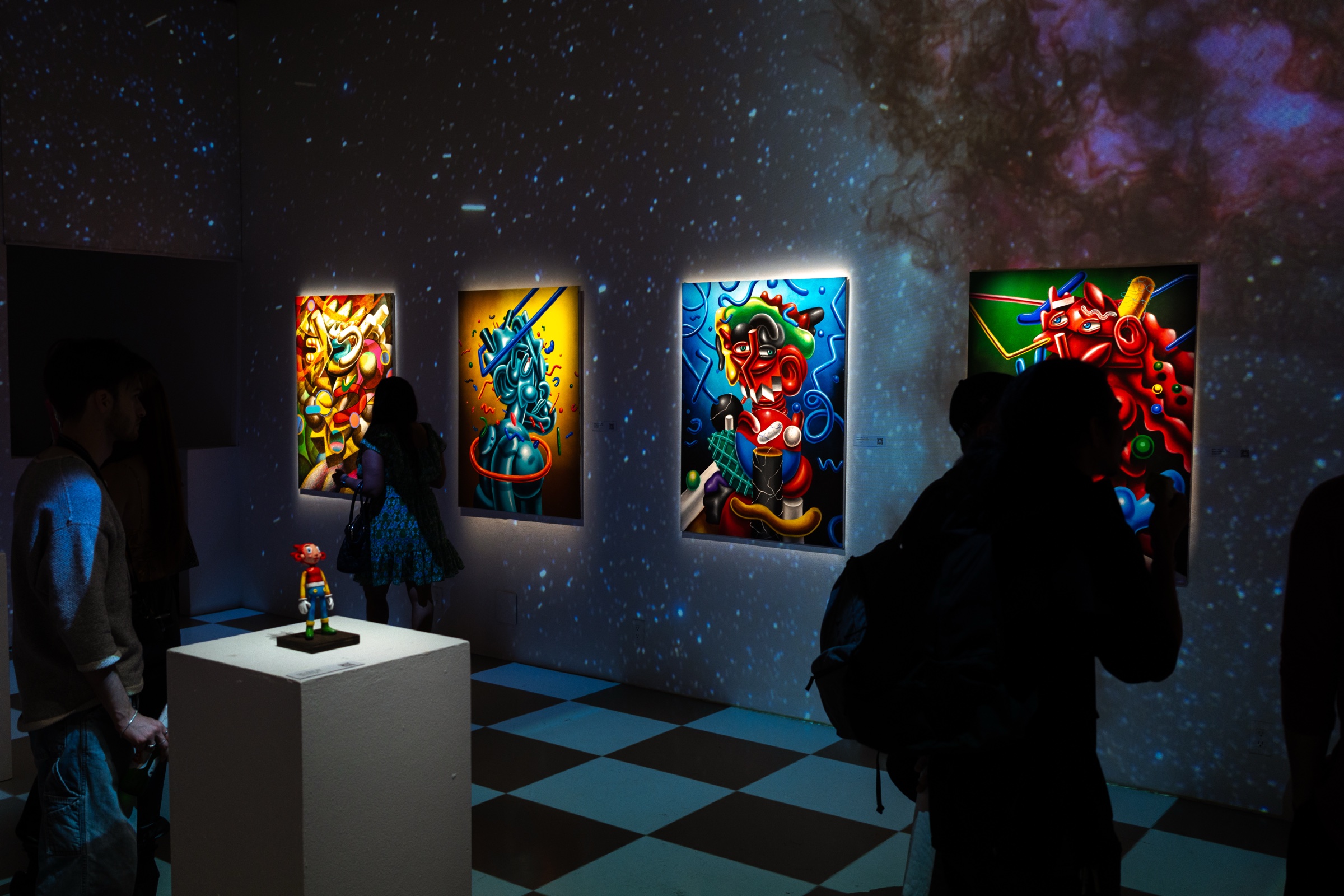Visitors in silhouette before four lit paintings, a small red figurine on a plinth at left