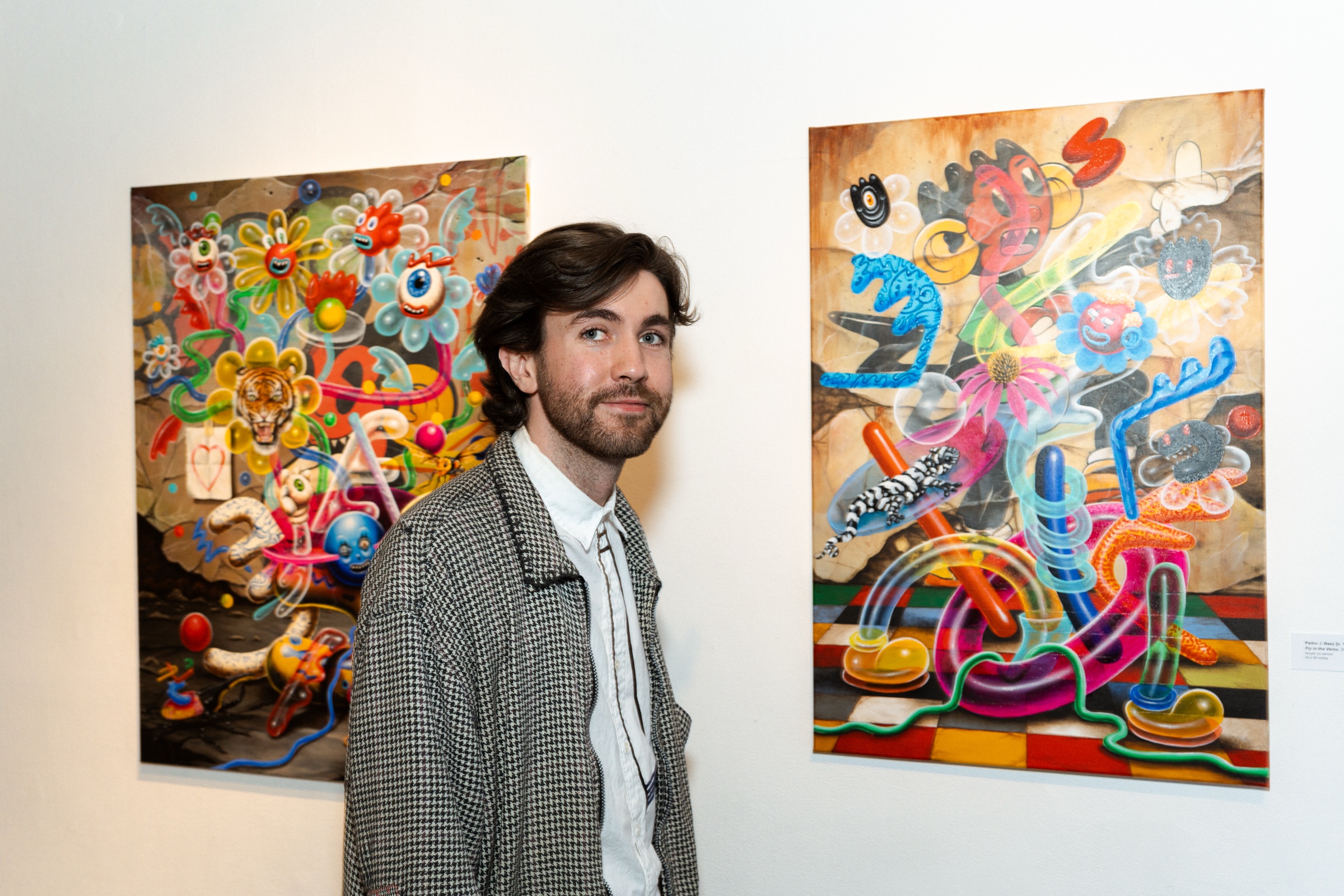 A man in a checked blazer stands between two paintings of tangled cartoon shapes on a white wall