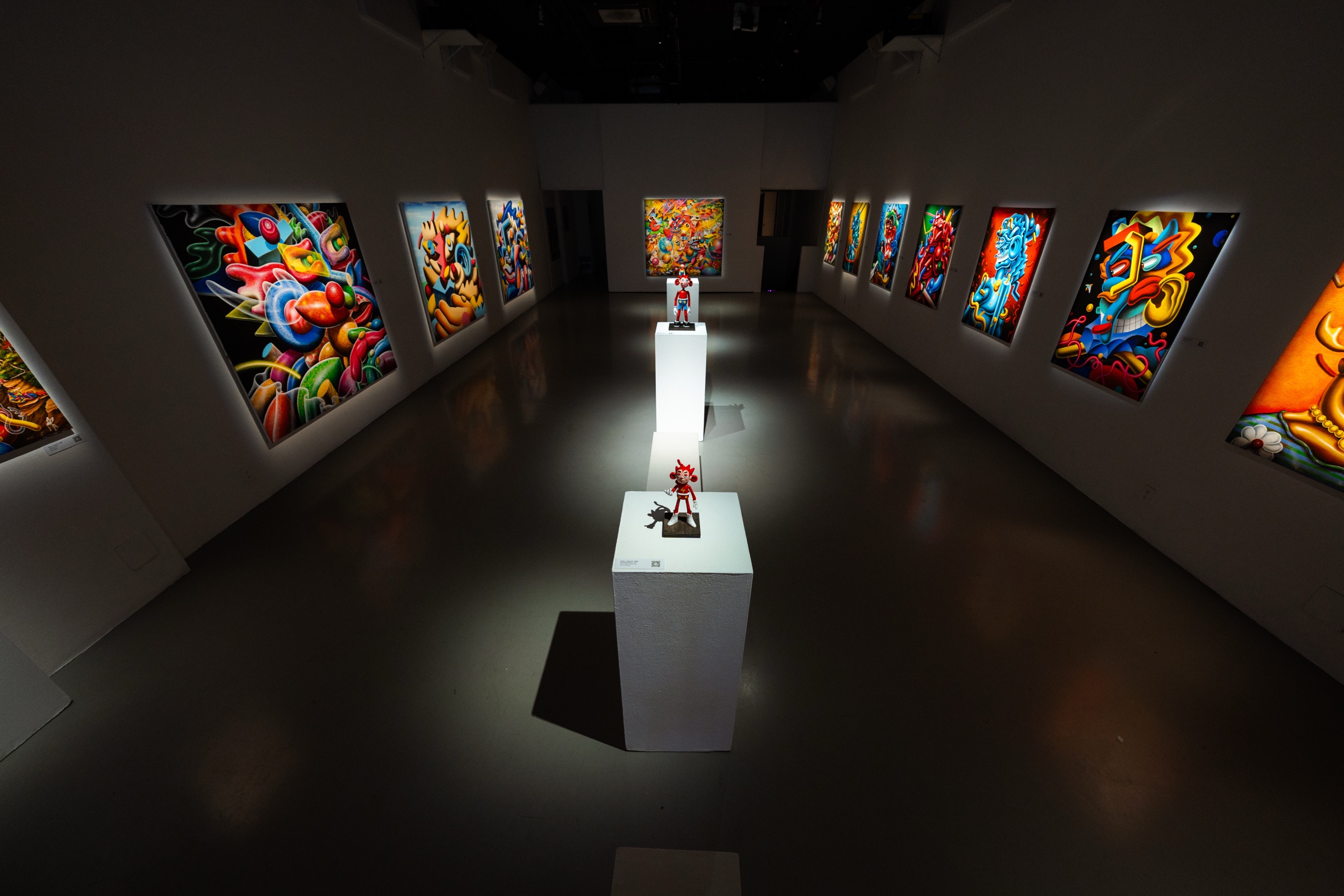 Darkened gallery with lit paintings down both walls and two small sculptures on white plinths in the centre