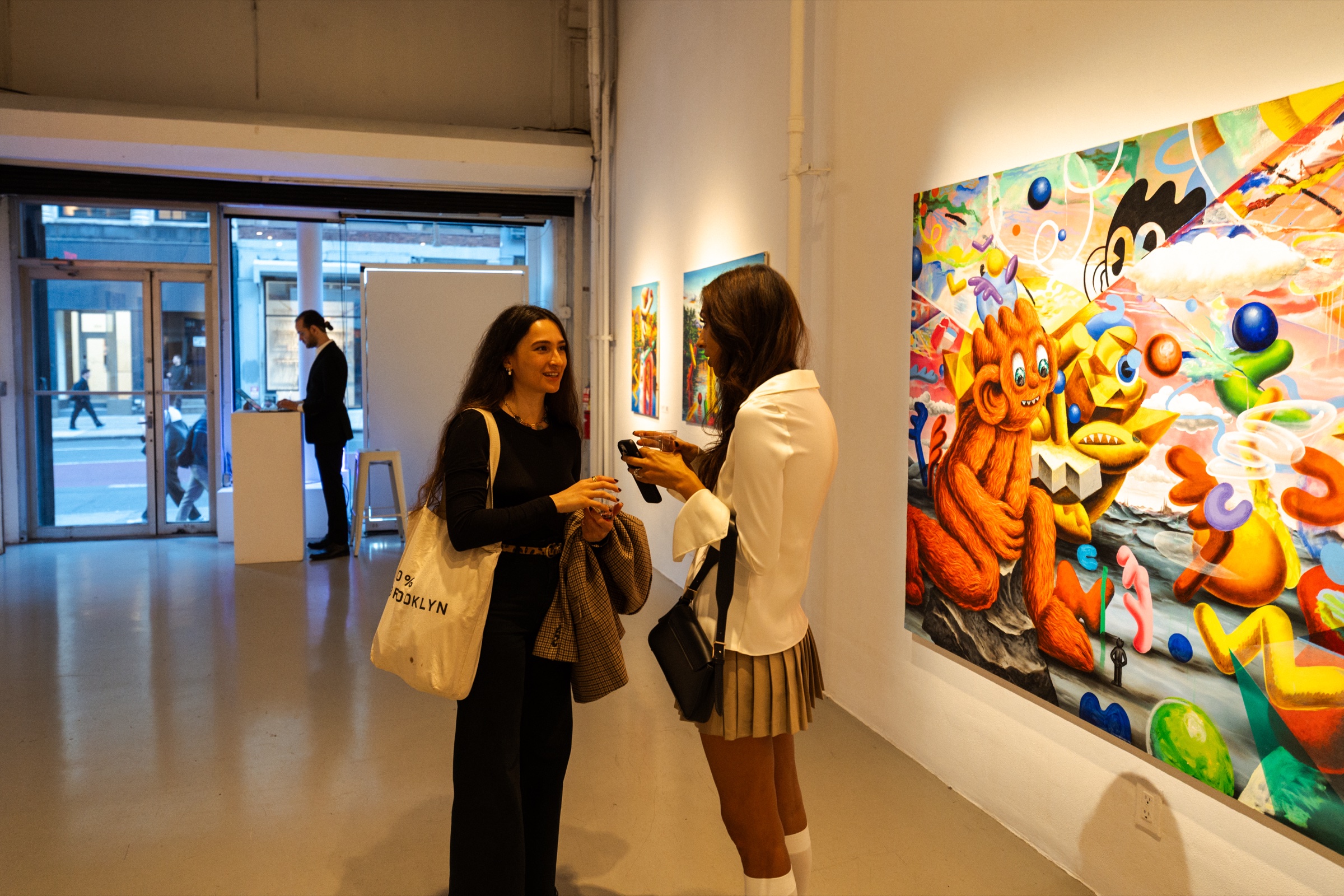 Two women talk beside a large painting of an orange creature near the gallery’s glass street doors