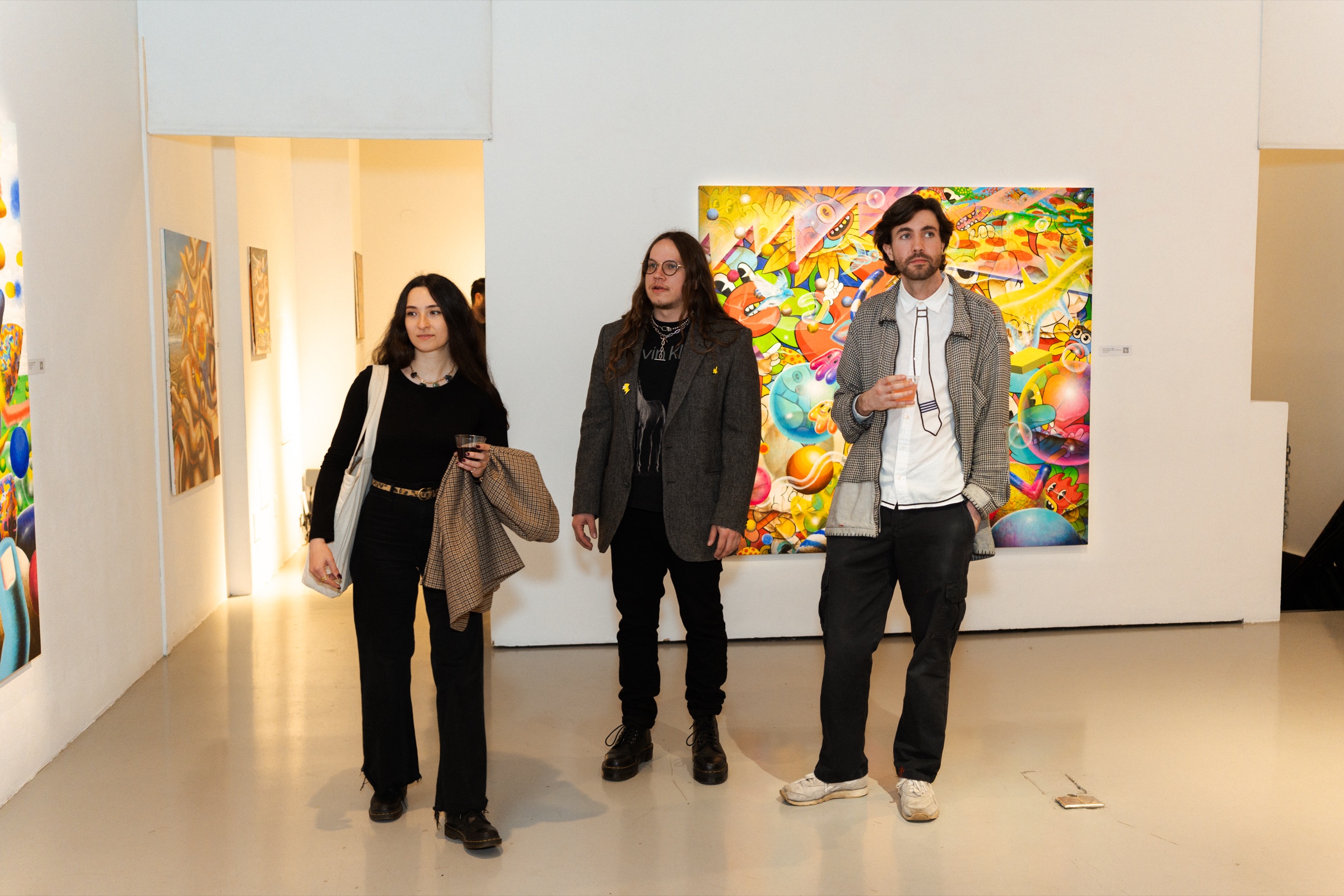 Three guests walk through the white gallery past a painting crowded with cartoon faces and balloons