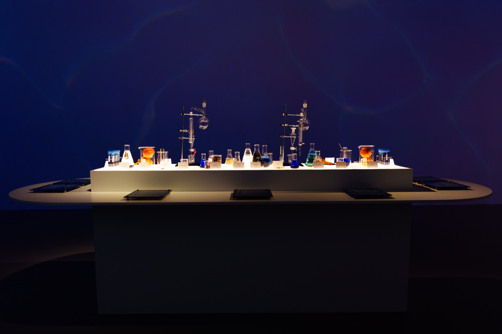 Glass lab flasks and beakers glow on a long white table in a dark blue room