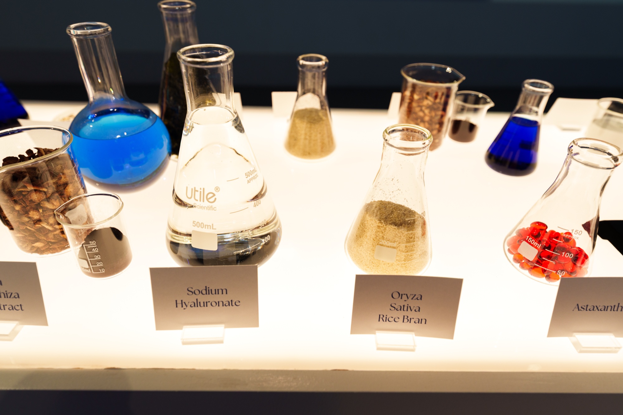 Glass flasks of blue liquid, rice bran, red capsules and wood chips labelled with ingredient names on a lit shelf