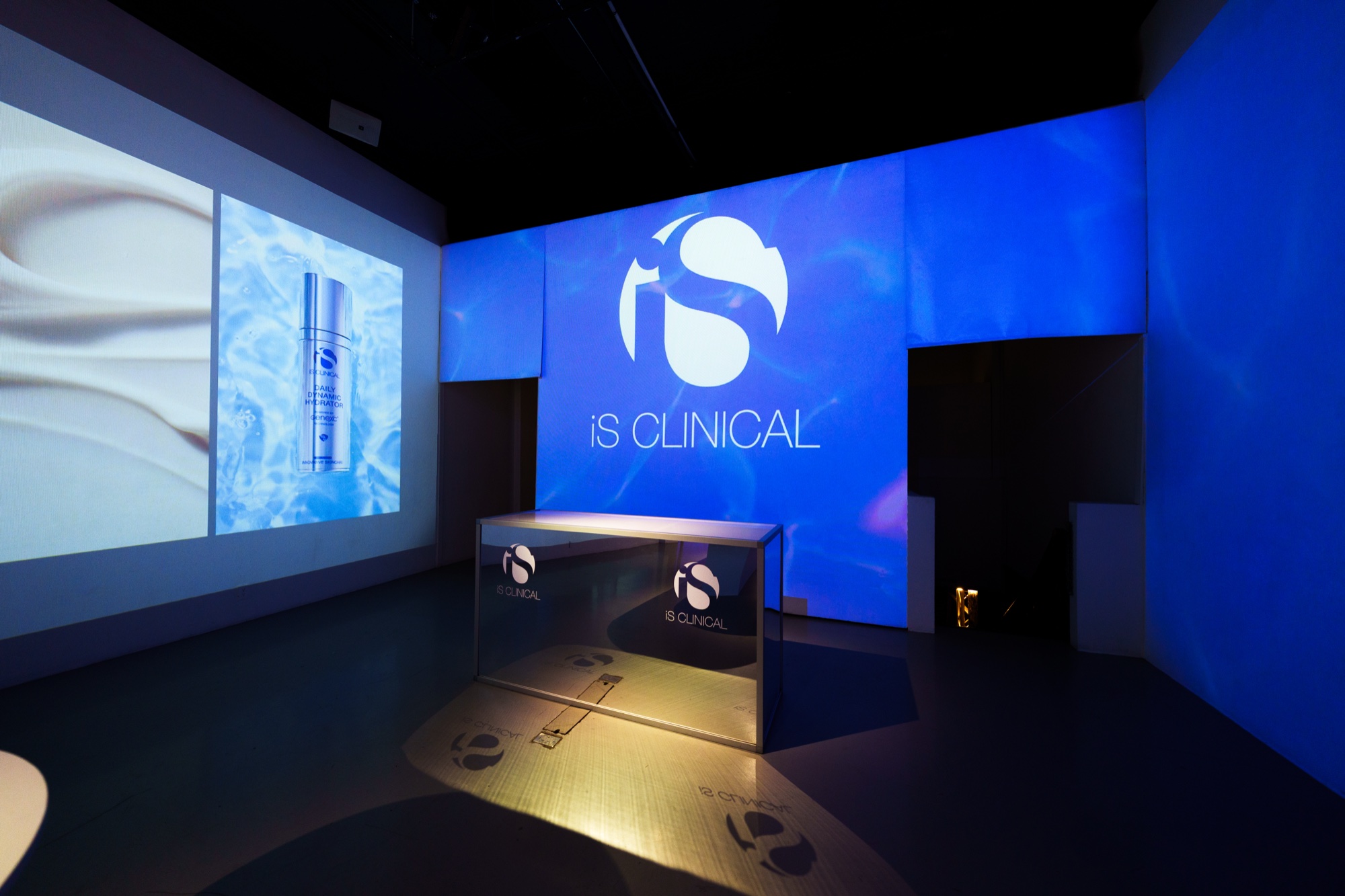 iS Clinical logo projected in white on a blue wall above a mirrored counter, a serum bottle projected at left