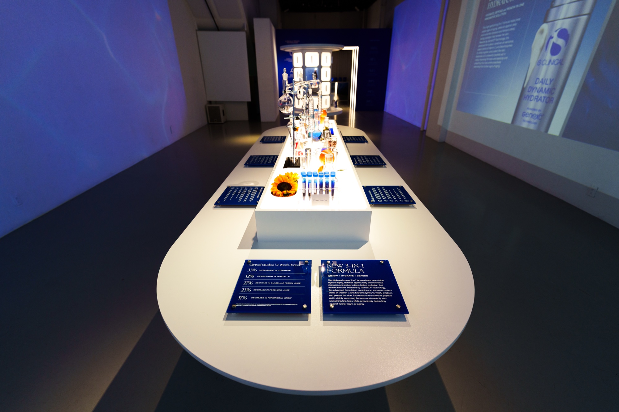 Long white oval table with dark blue info cards and lab glassware under a projected product ad