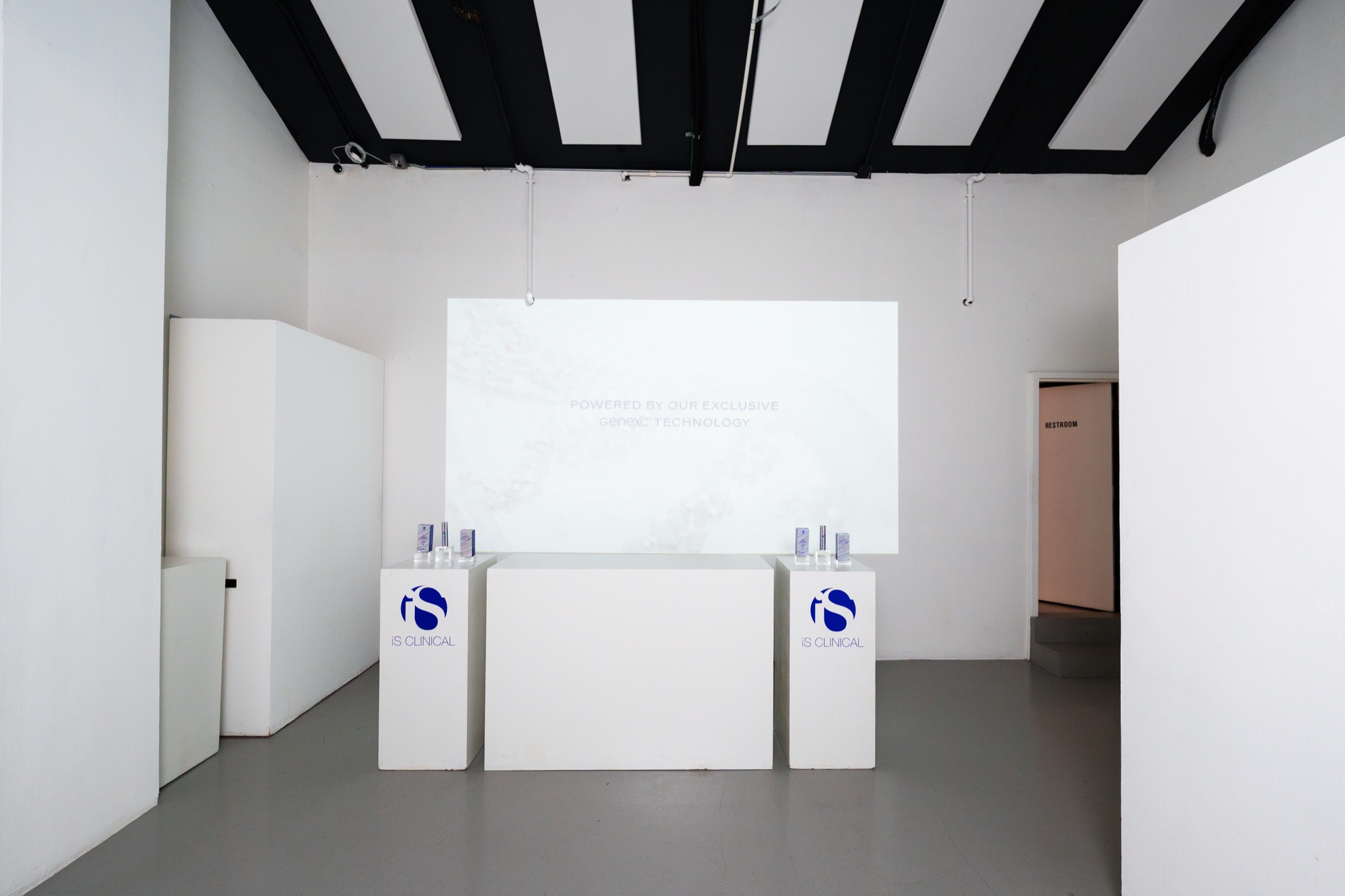White room with black ceiling beams, two iS Clinical plinths holding product bottles beside a blank projection screen