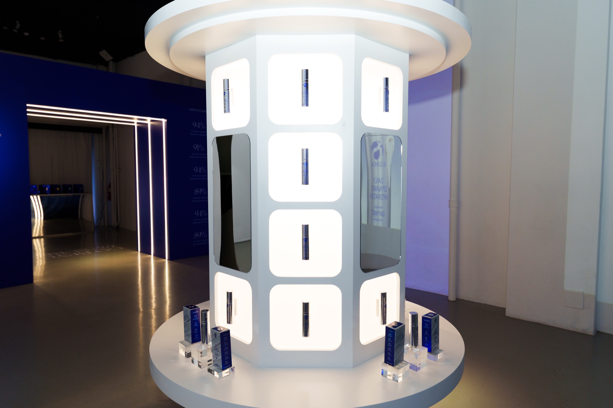 Round white column display of backlit panels holding serum bottles, blue lit corridor behind