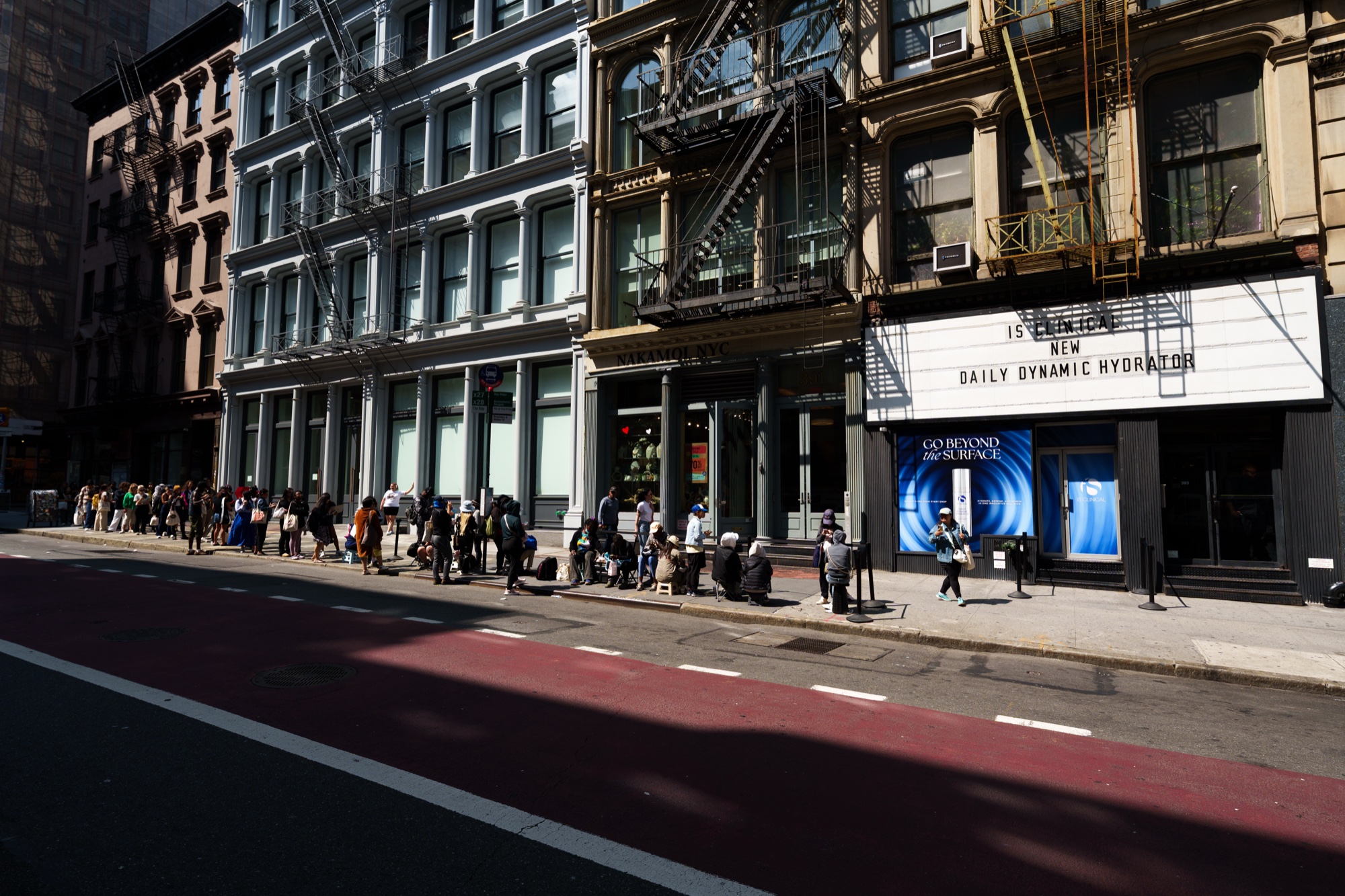 Line of people waits along a sunny city sidewalk beneath a marquee reading iS Clinical Daily Dynamic Hydrator