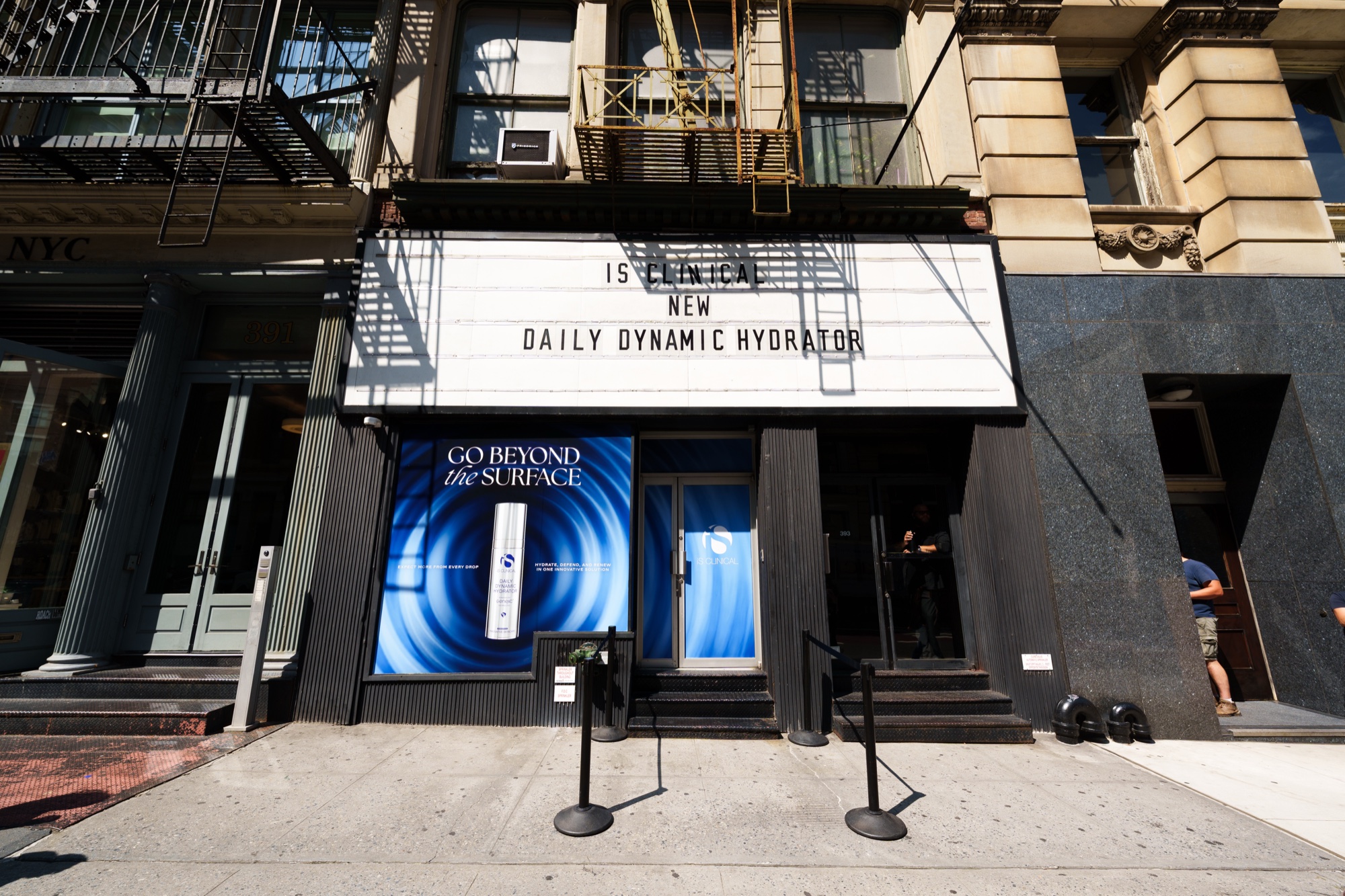 Storefront marquee reading iS Clinical New Daily Dynamic Hydrator, blue window graphics and a fire escape above