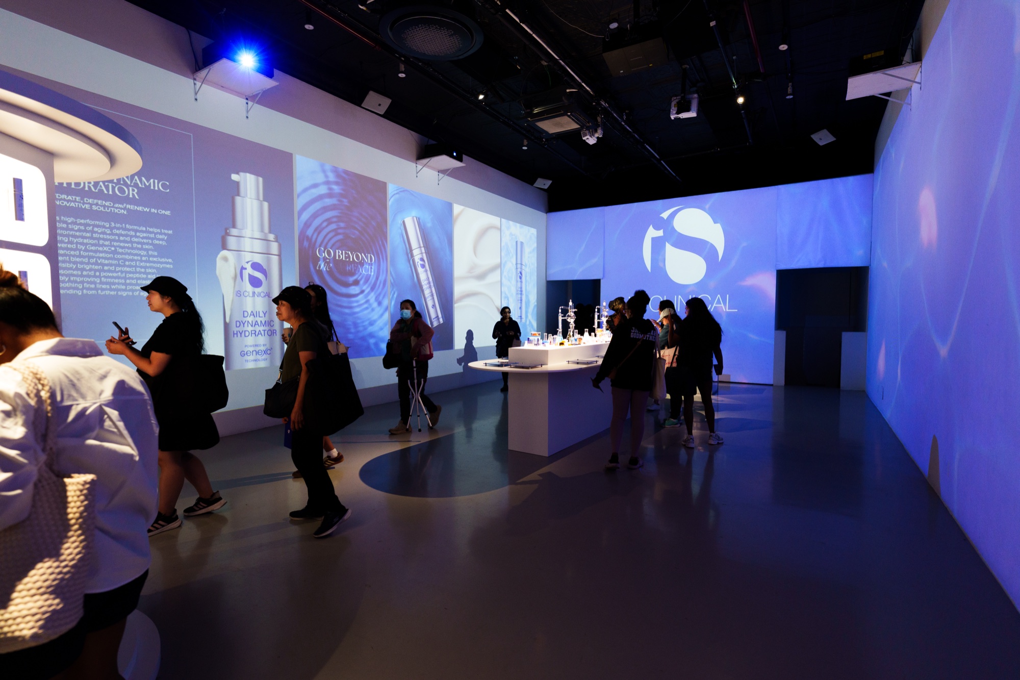 Visitors move through a blue lit room with projected serum bottles and an iS Clinical logo on the walls